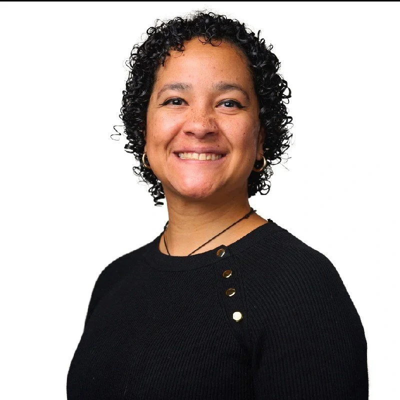 A woman with short, curly black hair and brown skin, smiling, wearing a black top with gold button details and gold hoop earrings, against a white background.