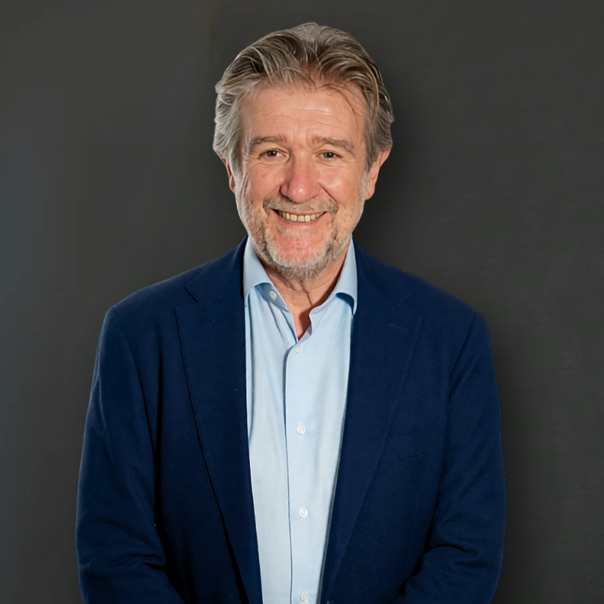 Close-up of a smiling middle-aged man with gray hair and a beard, wearing a suit jacket and dress shirt, outdoors with greenery and a white support structure in the background.