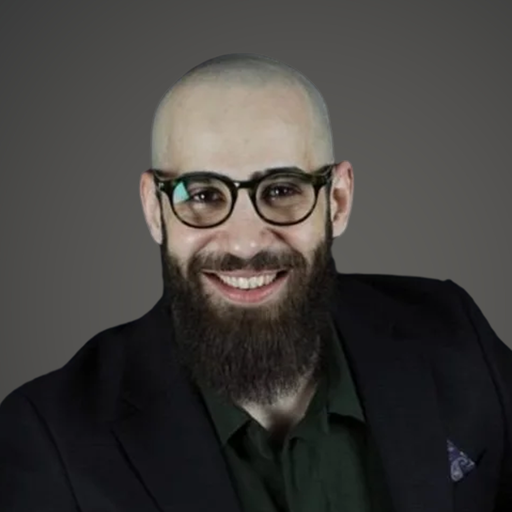 A man with a shaved head, beard, glasses, wearing a dark jacket and green shirt, smiling against a dark background.