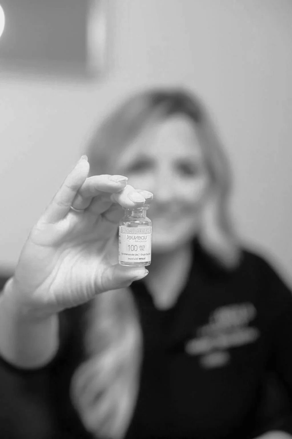 Person wearing black gloves holding a syringe and vaccine vial, preparing an injection, with a tattooed arm visible, in a clinical setting.