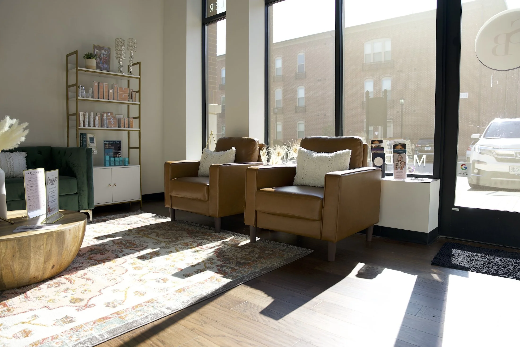 Modern waiting room with brown leather chairs, a wooden coffee table, decorative shelf with products, large windows, and a patterned rug.