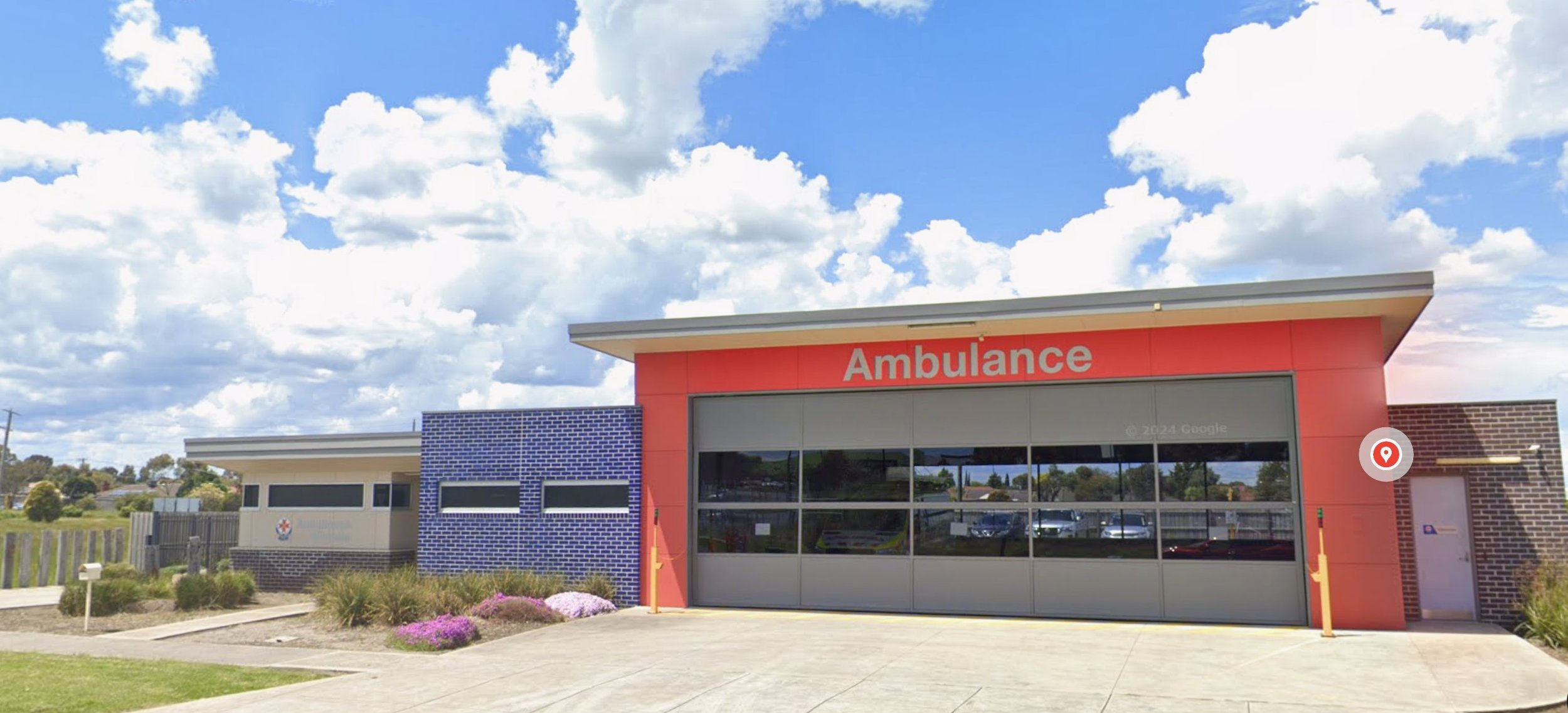 A modern ambulance station with a red sign reading 'Ambulance' on the building, a large garage door, and blue and brick exterior walls, under a partly cloudy sky.