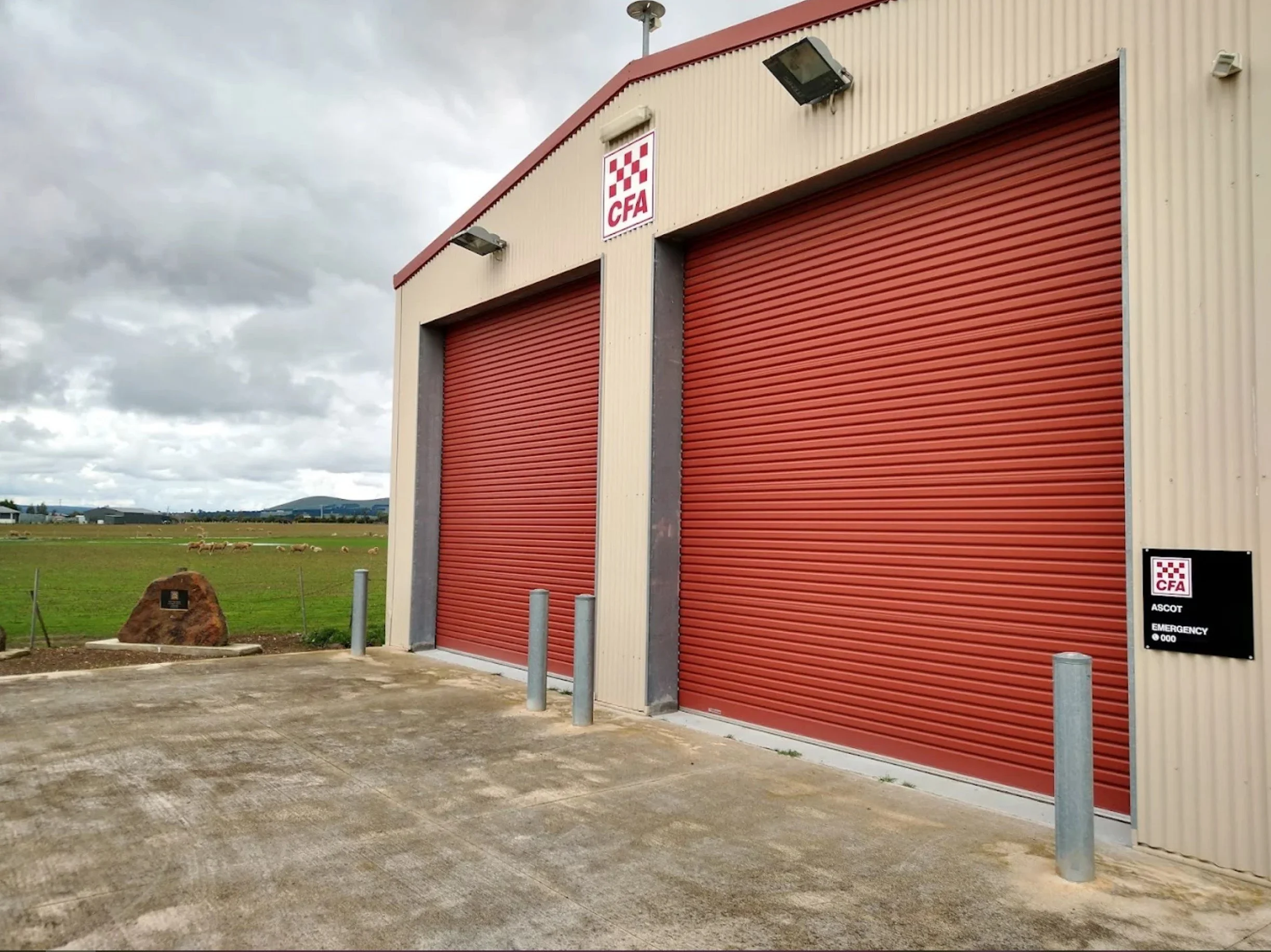 Agricultural building with red roll-up doors and CFA logo, with a grassy field and sheep in the background.