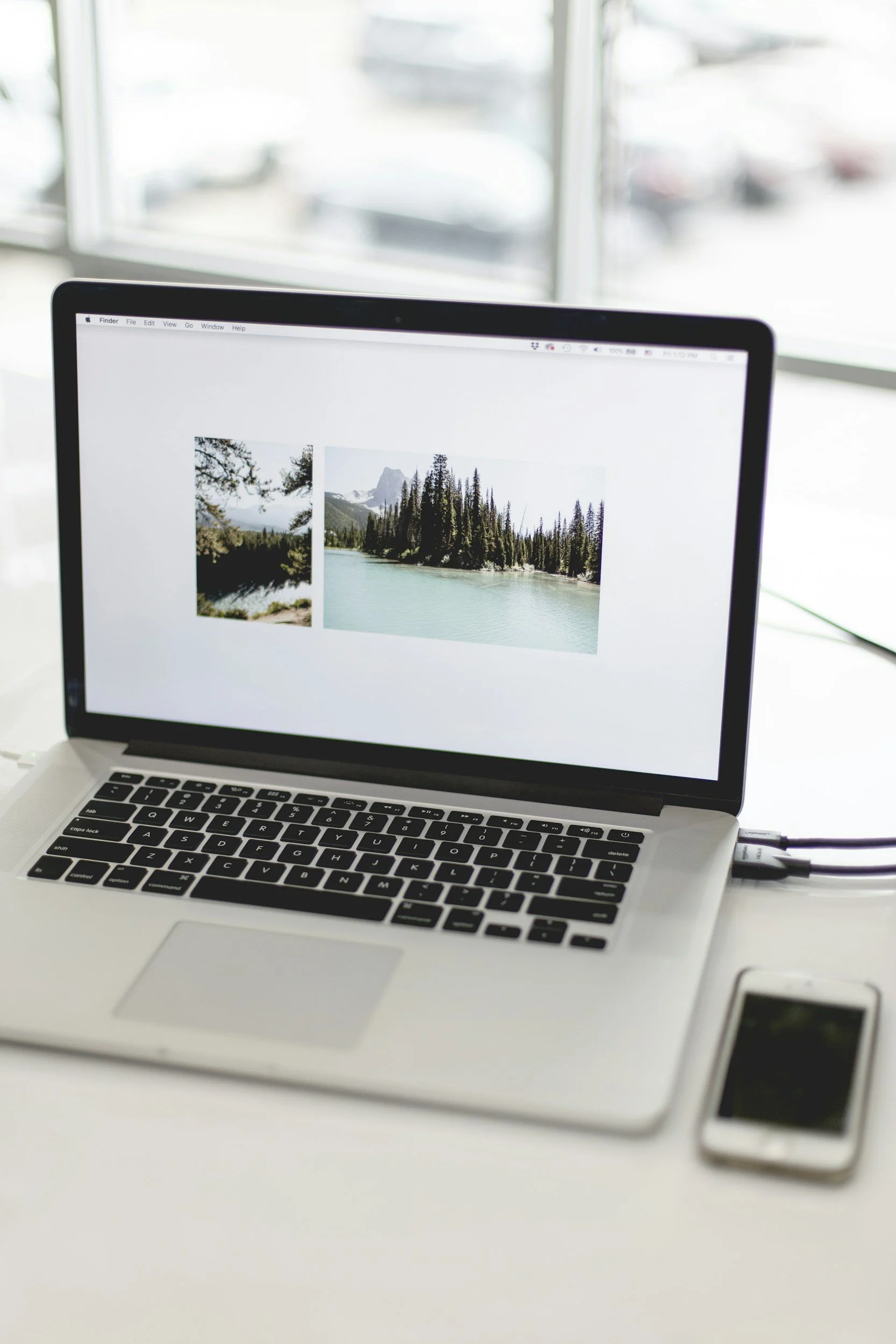 Laptop on a white desk with a screen displaying images of a lakeside landscape with trees and mountains, and a smartphone lying beside it.