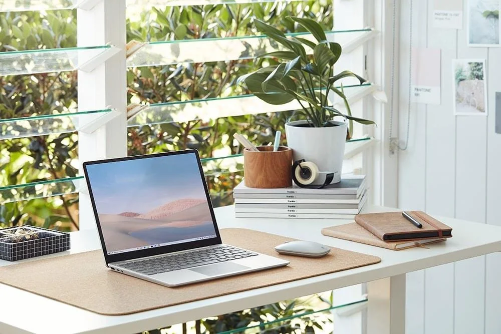 virtual assistant Home office desk with a laptop, mouse, notebooks, a plant, and books near a window with greenery outside.