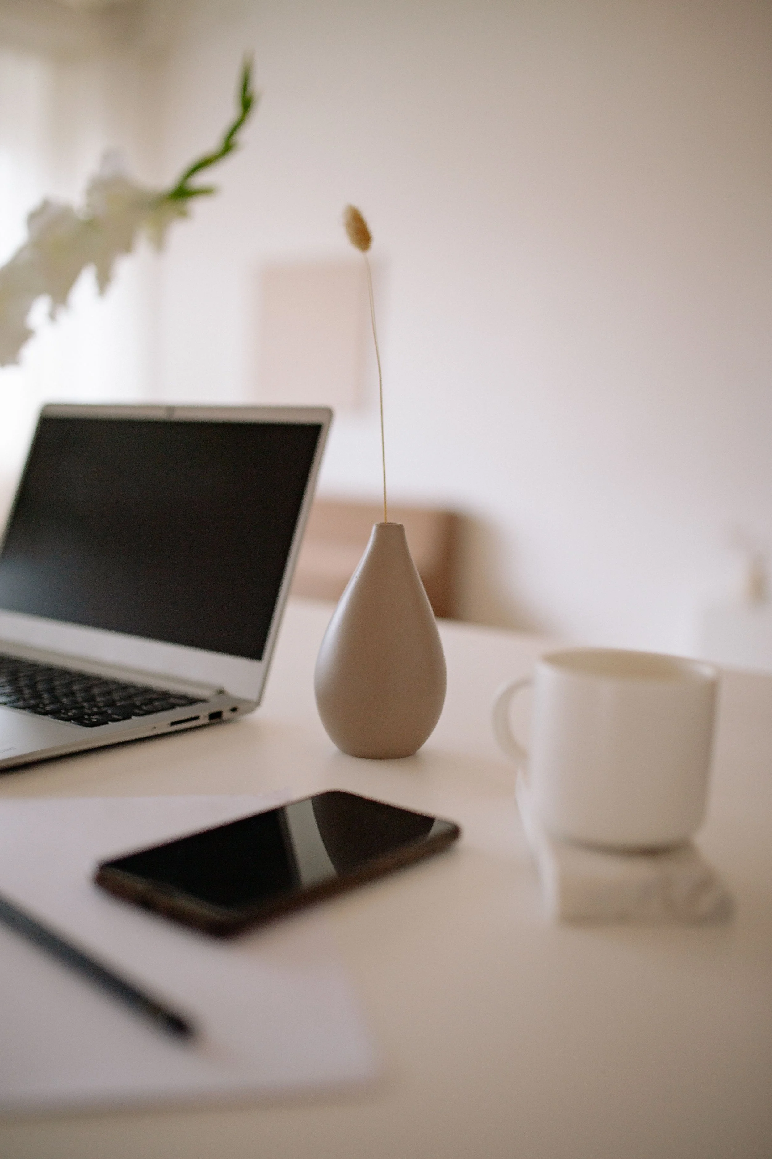 A virtual assistant minimalist workspace with a laptop, a smartphone, a white mug, and decorative items including flowers in a vase and a ceramic object on a white desk.