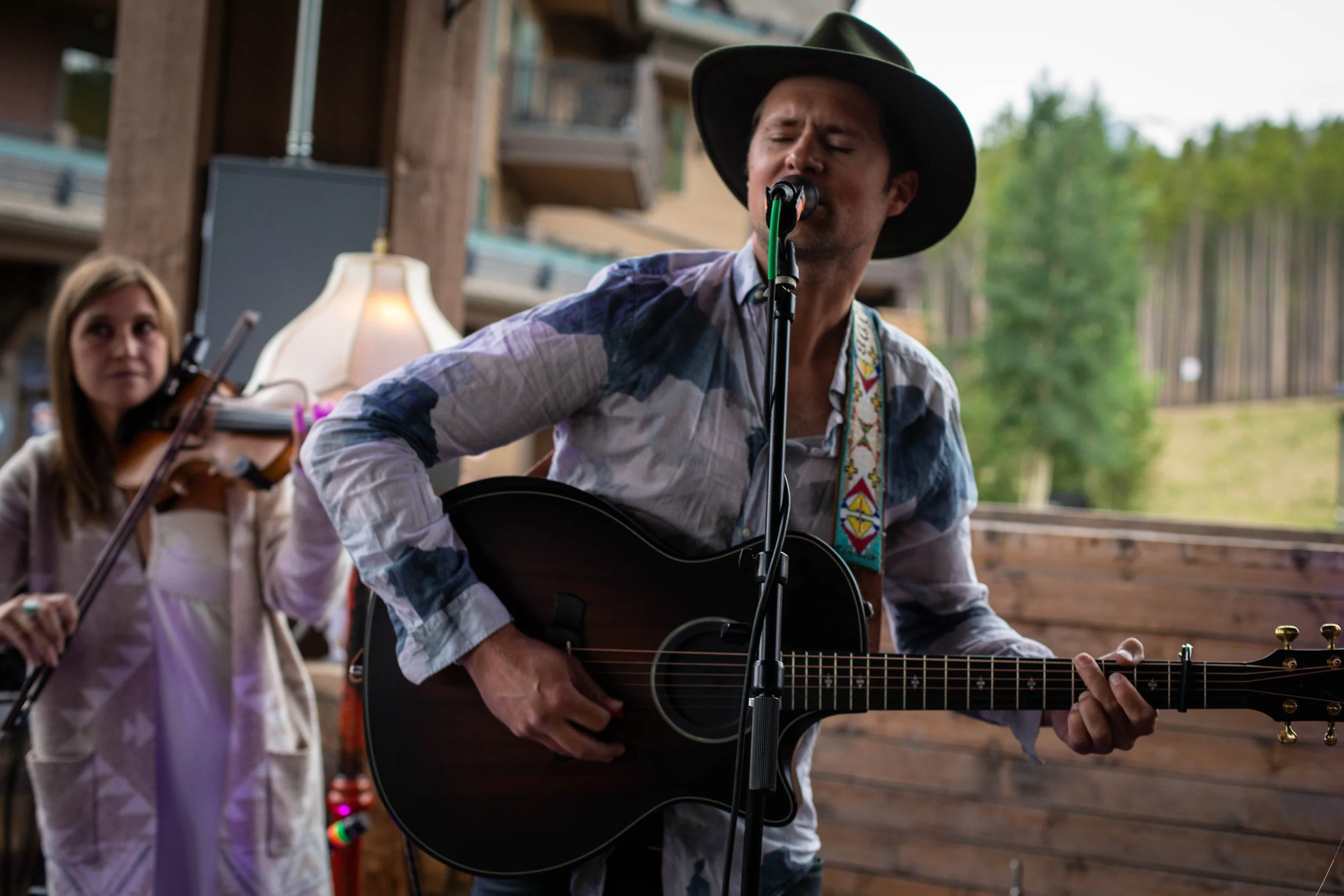 A man wearing a cowboy hat and a patterned shirt playing an acoustic guitar and singing into a microphone. In the background, a woman with a violin. The setting appears to be an outdoor or semi-outdoor rustic venue with wooden walls and greenery outs