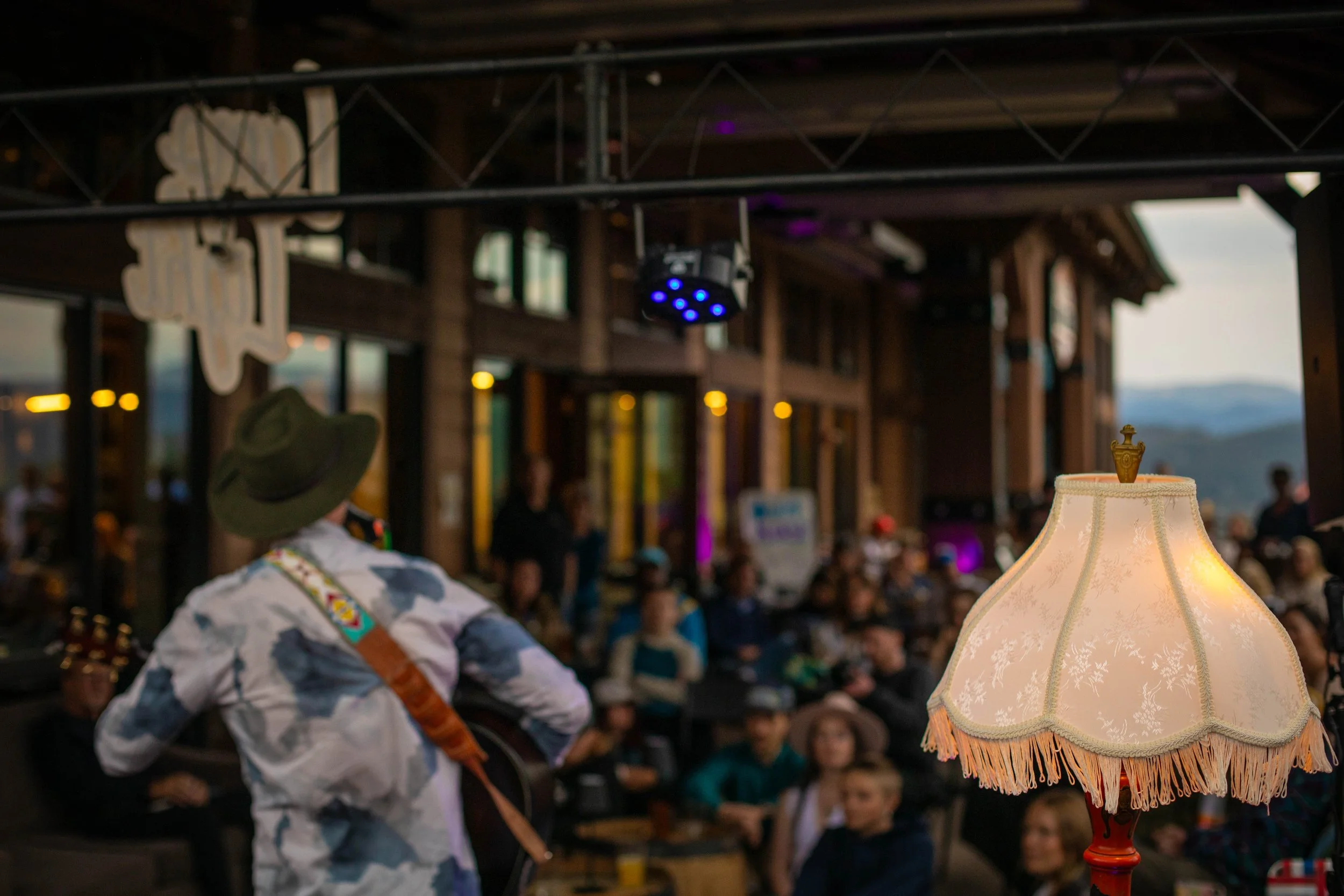 A musician wearing a hat and camouflage shirt performing with a guitar at an outdoor venue during the evening, a vintage-style lampshade in the foreground, and an audience watching the performance.