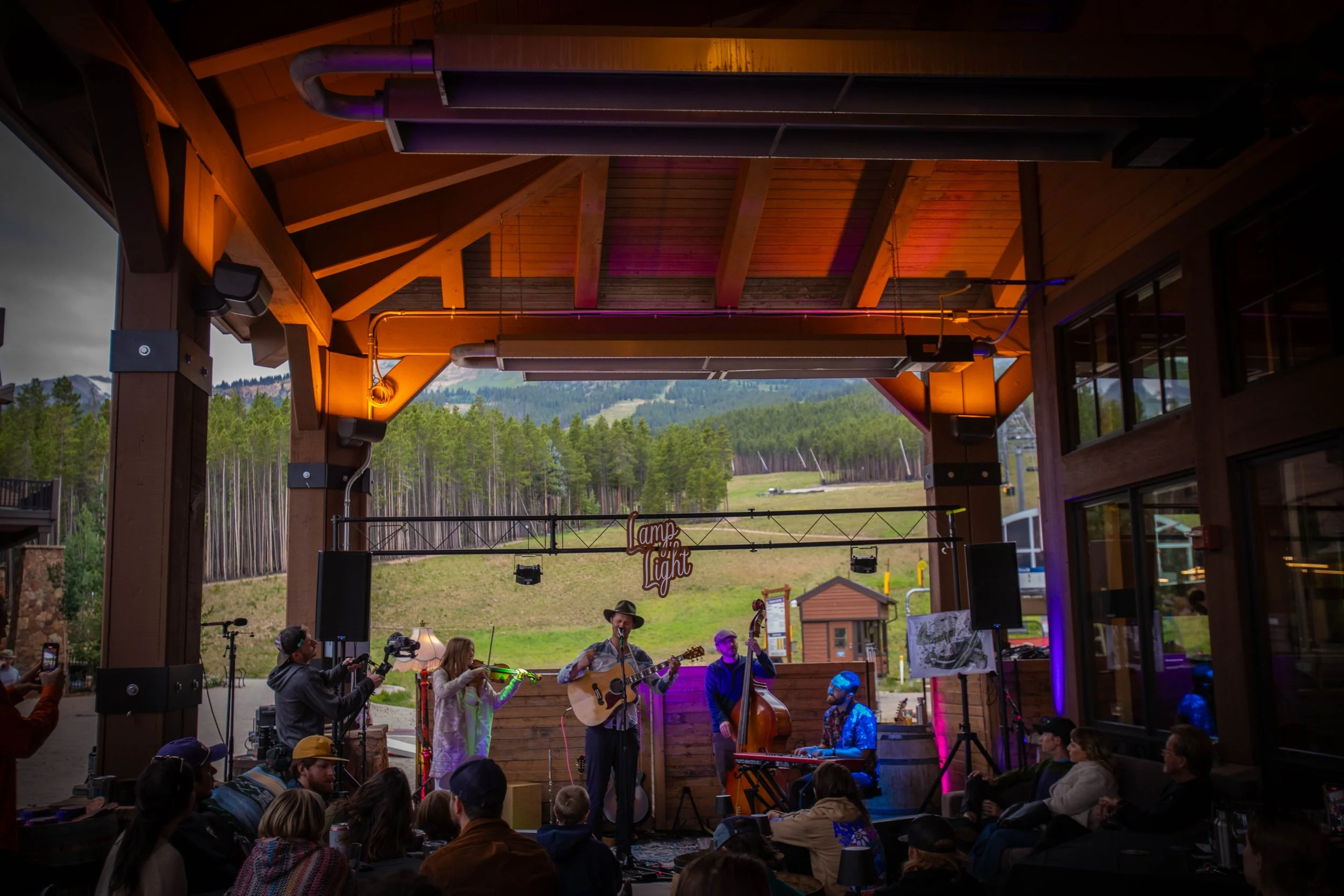 Band performing on an indoor stage with an outdoor mountain landscape in the background, audience seated watching, and a sign that says "camp light".