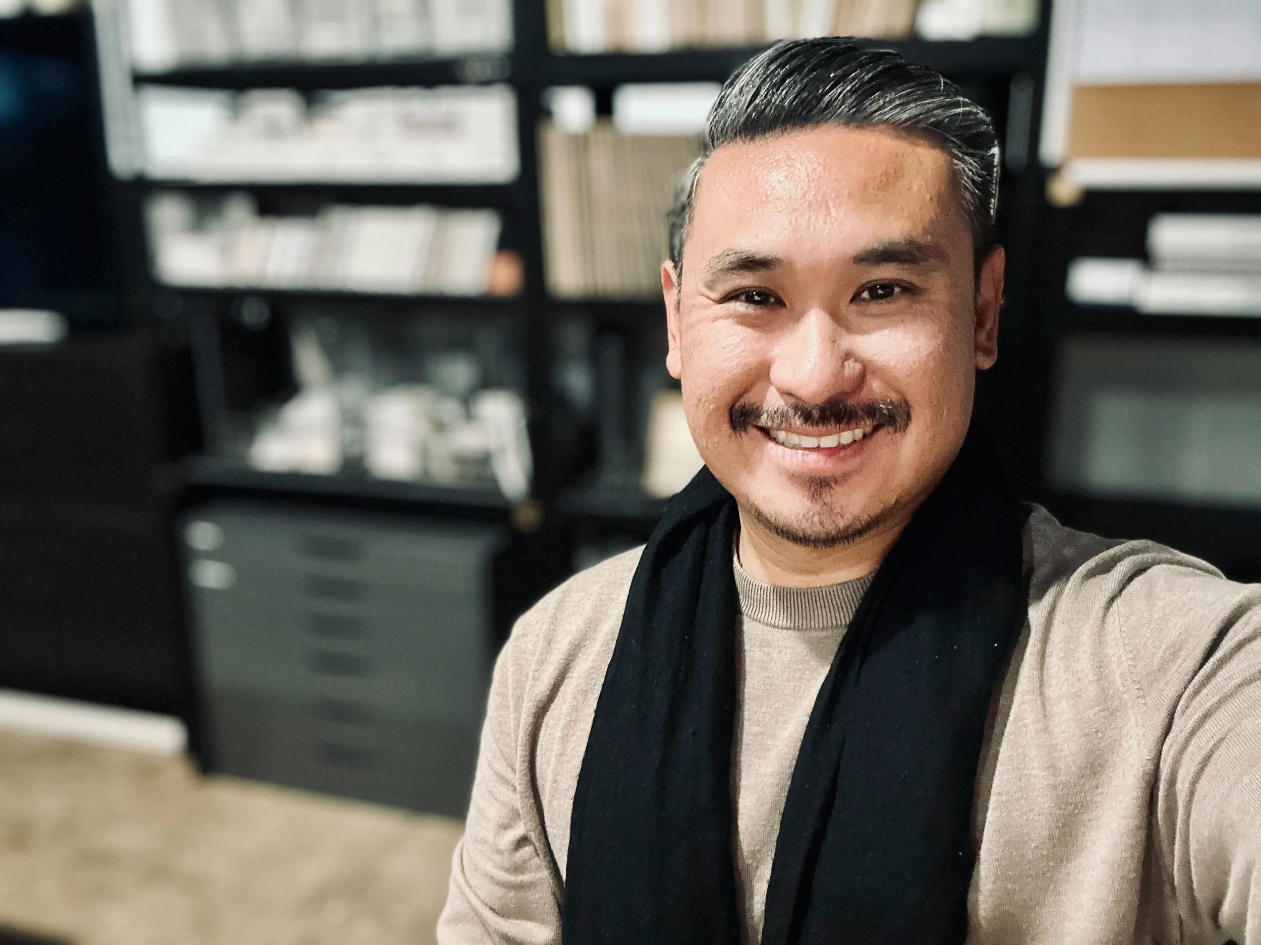 Smiling man with short dark hair and facial hair, wearing a beige sweater and a black scarf, taking a selfie indoors with a background of bookshelves filled with books.