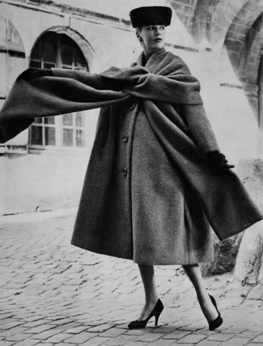 A black-and-white photograph of a woman wearing a large hat and a long, elegant coat, walking on cobblestone pavement with an arched stone bridge in the background.