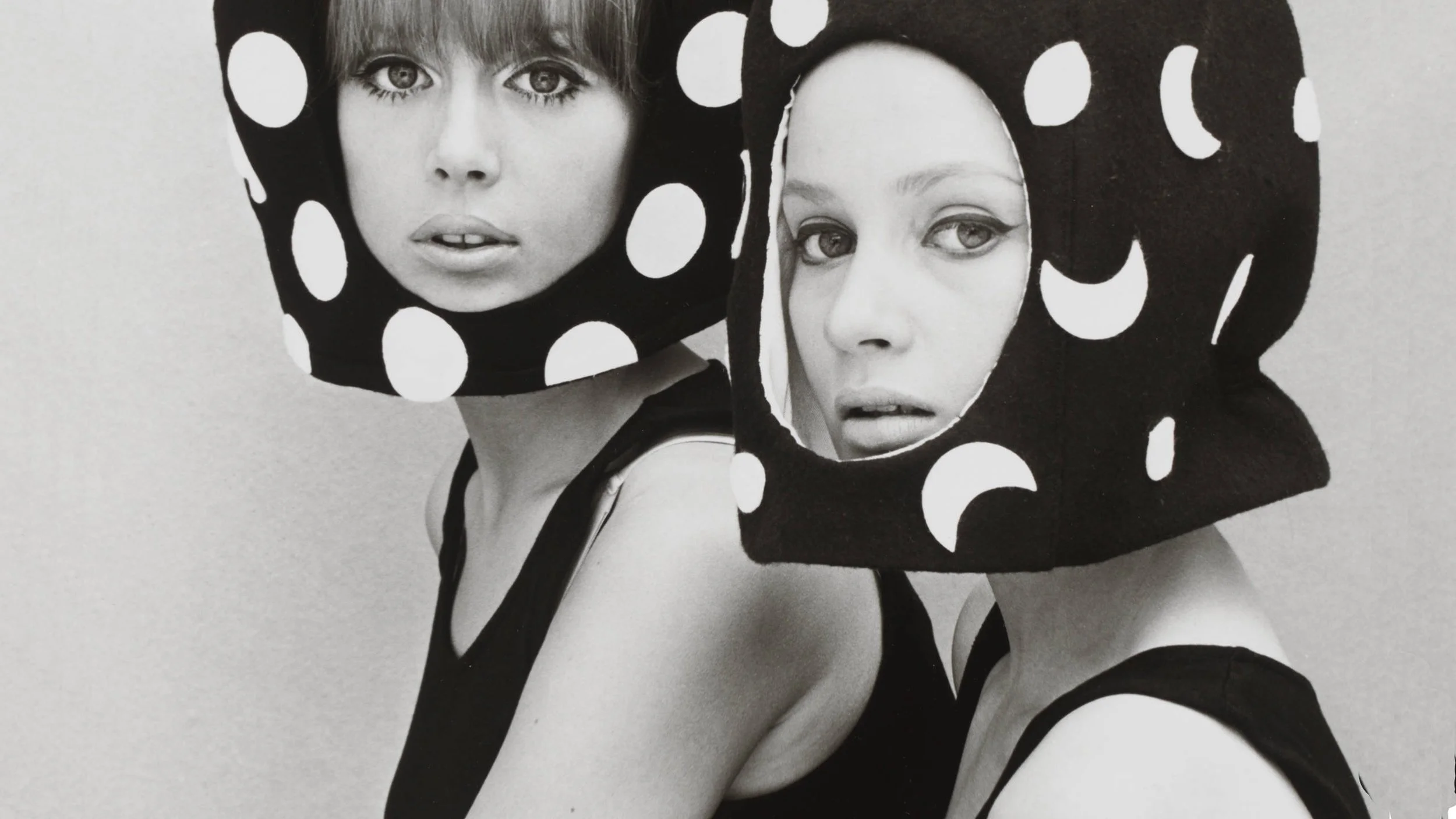 Two women wearing black headpieces with large white polka dots and cut-out faces, posing against a plain background