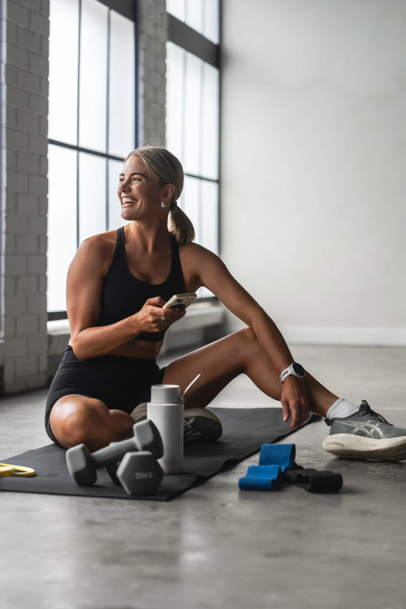 A woman sitting on an exercise mat in a bright fitness studio, smiling and looking away, holding a phone with a netball program on screen. She has workout gear, a smartwatch, and a water bottle nearby, with exercise equipment around her.