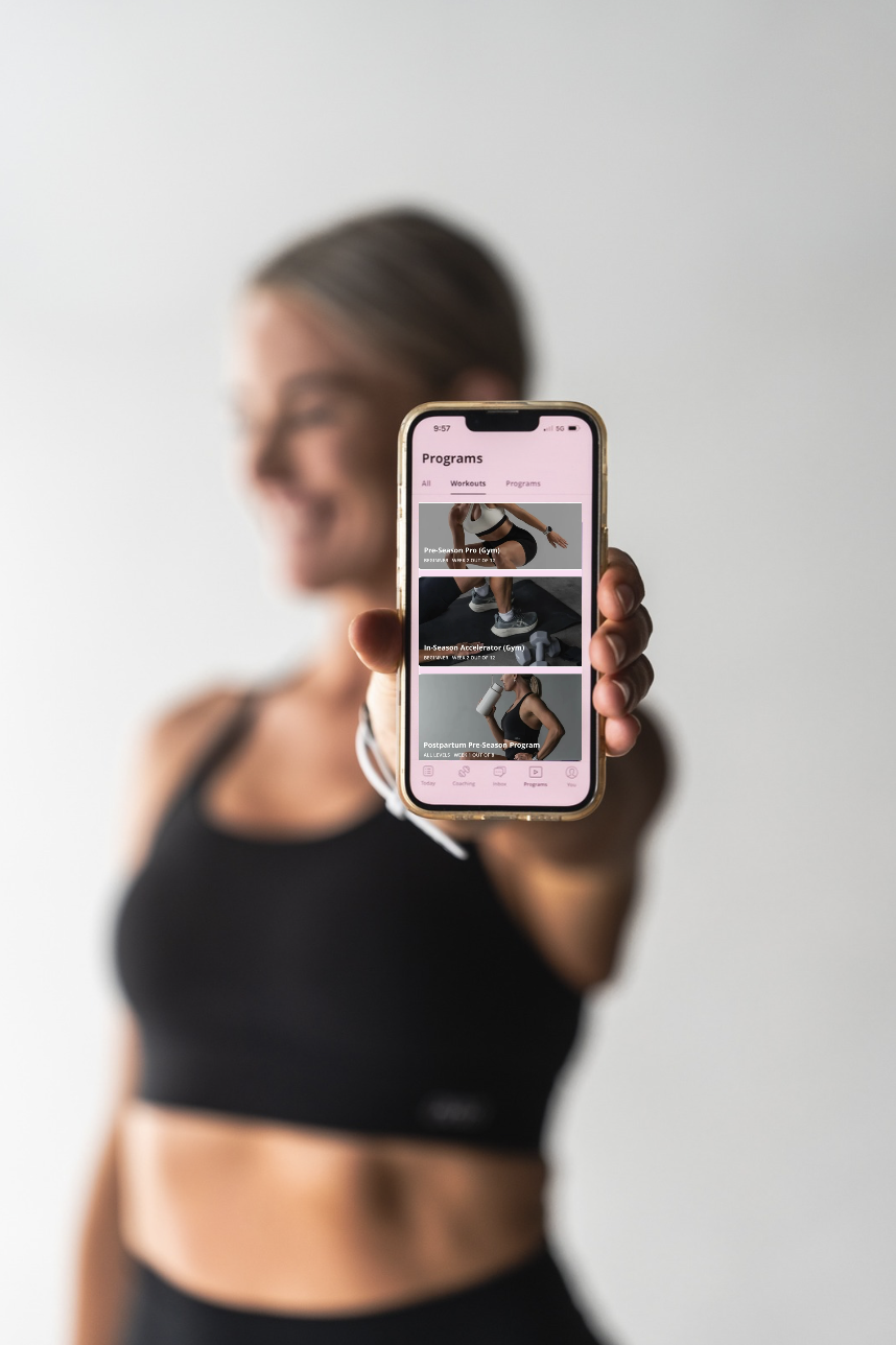 Blurred woman in athletic wear holding a smartphone displaying netball strength & conditioning programs on a pink app interface.
