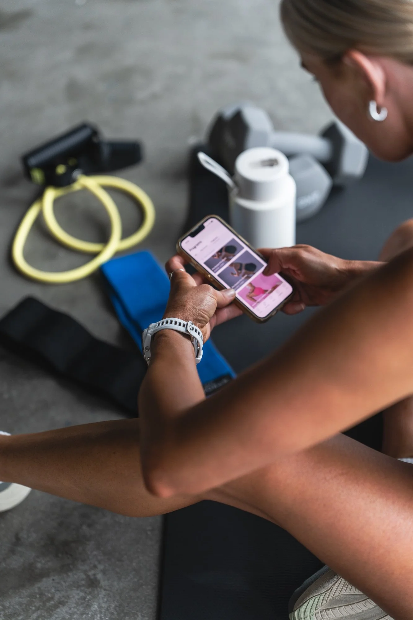 Person sitting on the floor in a gym, looking at a phone with netball strength and conditioning program. Gym equipment including dumbbells, resistance bands, a water bottle, and a yoga mat are nearby.