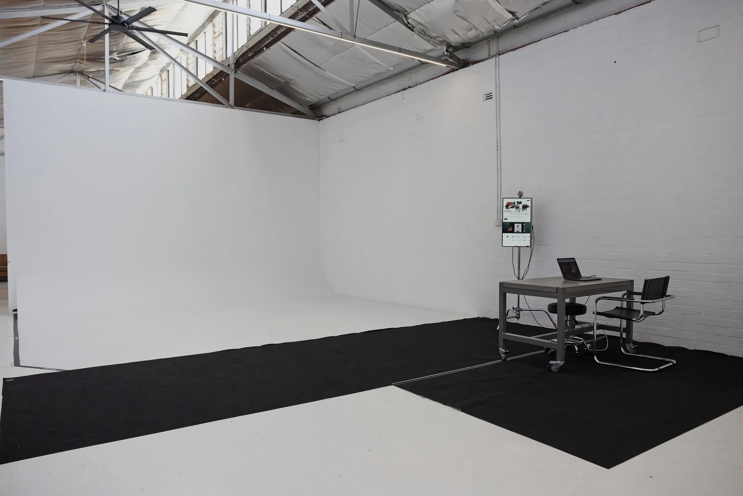 Empty photo studio with white wall and a black carpet, a small table with two chairs, and a mounted monitor on the wall.