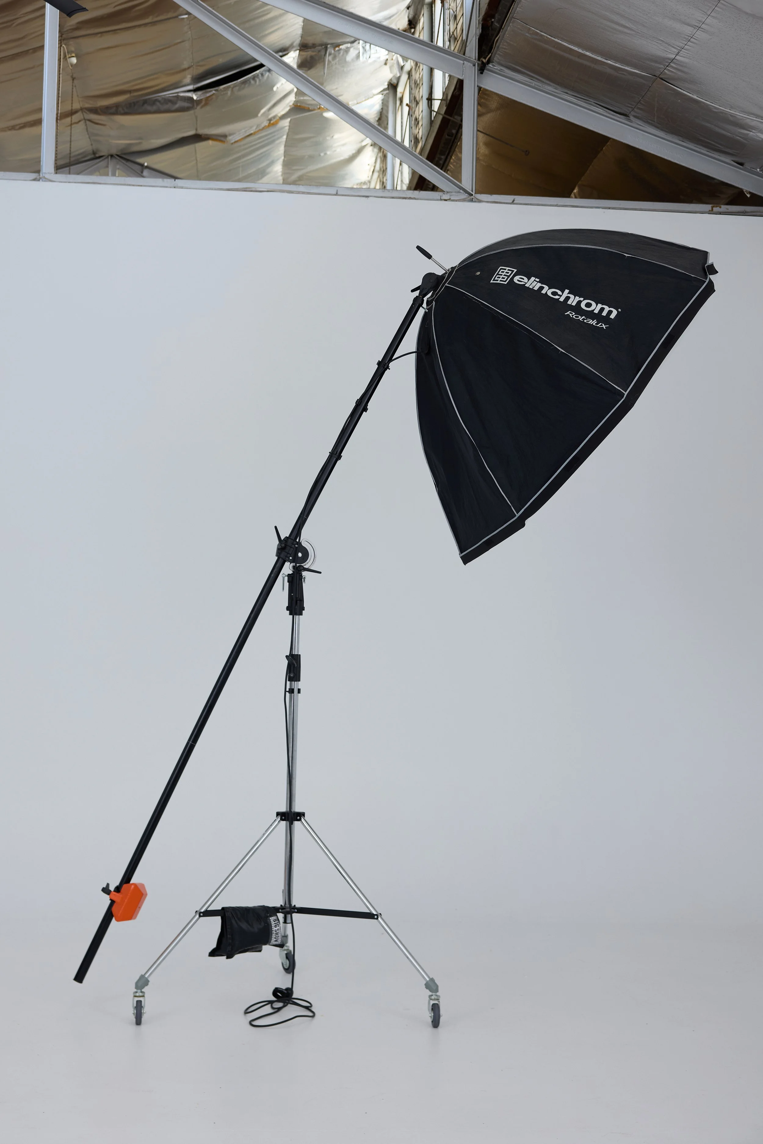 A photography studio setup with a black Elinchrom umbrella light modifier on a stand, positioned against a white backdrop, and a ceiling with metallic insulation and structural beams overhead.