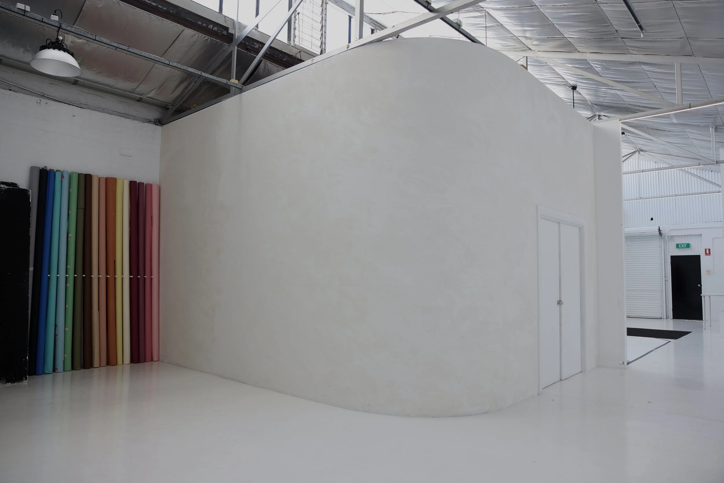 An interior space with a white curved wall, colorful poles stacked against a black surface, and an industrial ceiling with exposed metal beams and lighting.