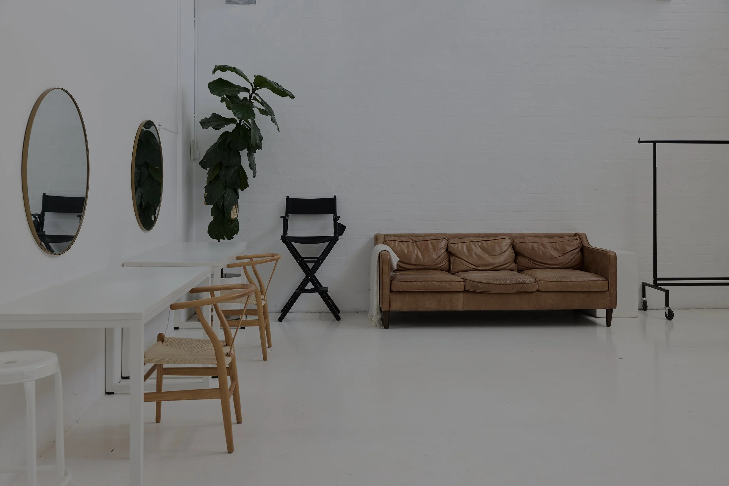 Minimalistic interior with white walls, a brown leather sofa, black folding chair, tall green plant, two round mirrors on the wall, a white table, several wooden chairs, a small white stool, and a black clothing rack on wheels.