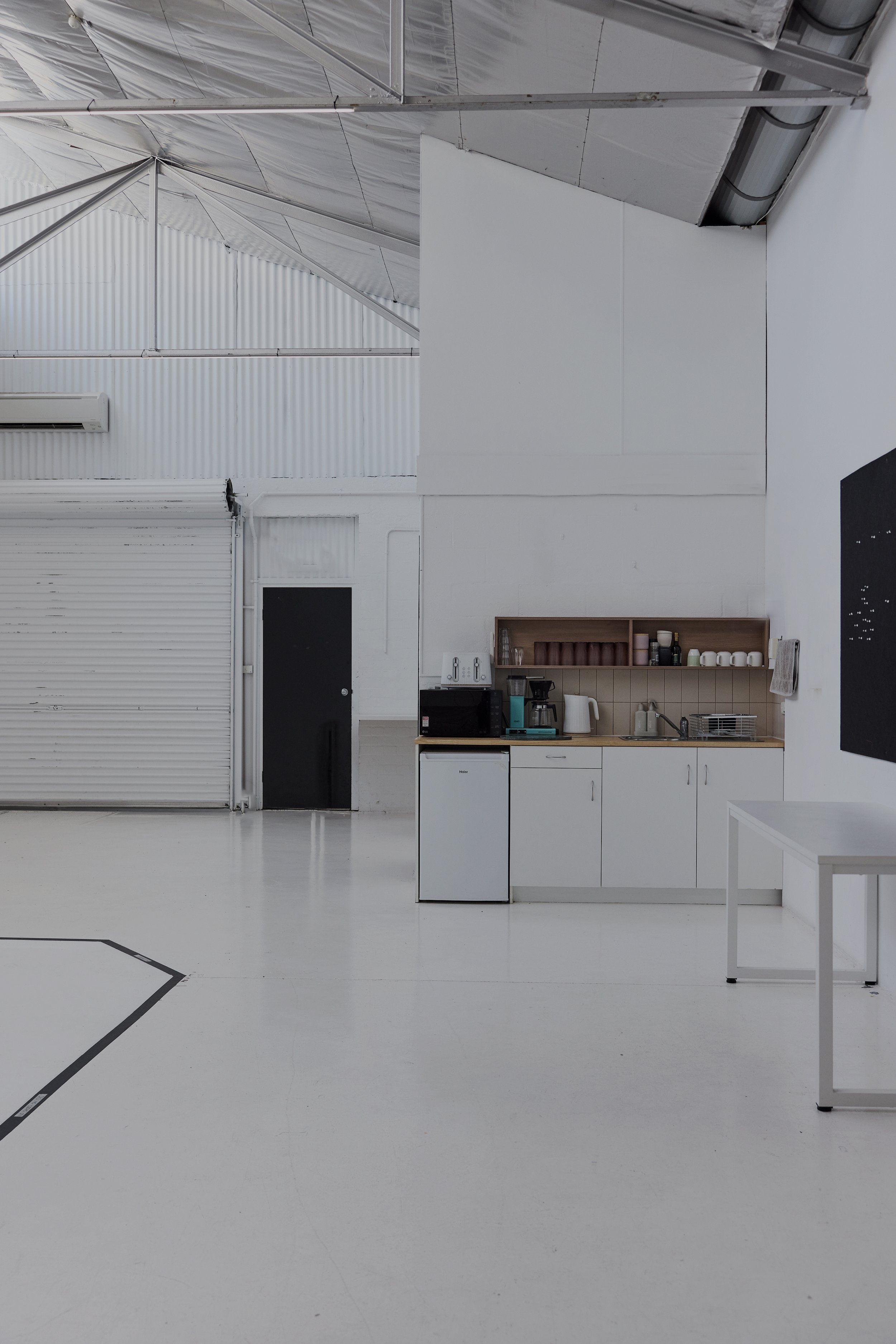 Minimalist kitchen setup inside a large industrial space with white walls, a small refrigerator, and shelves with cups and kitchenware.