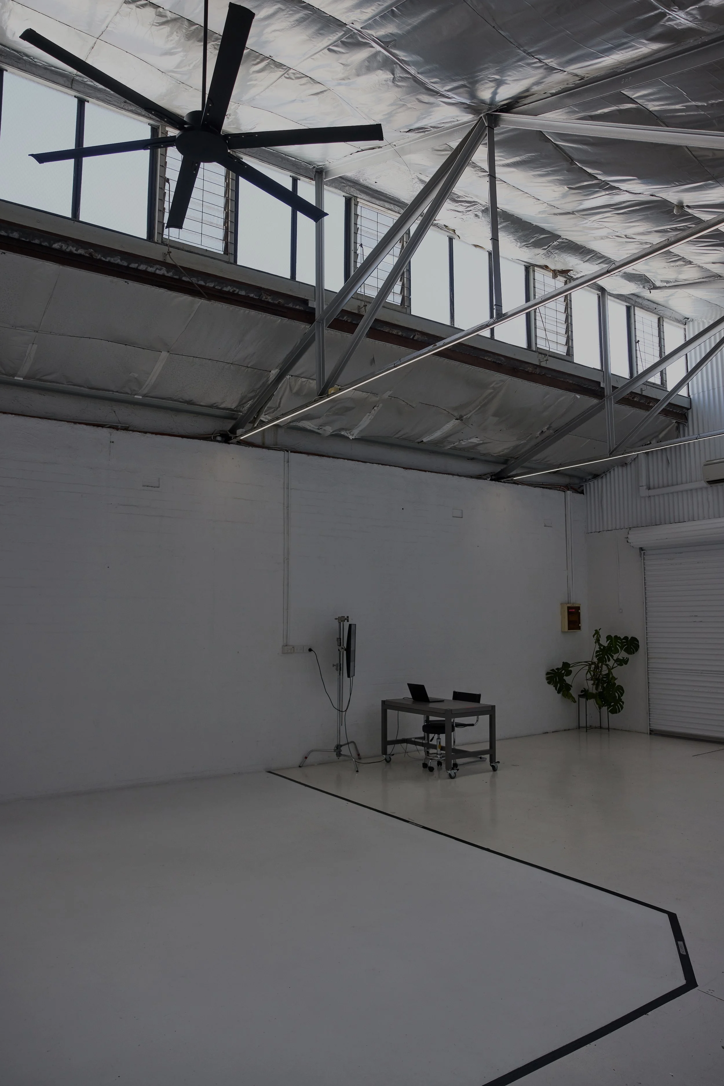 Interior of a spacious, industrial-style room with white walls, a high ceiling with exposed insulation, and a large ceiling fan. A small desk with a laptop and chair is against the wall, with a potted plant nearby.
