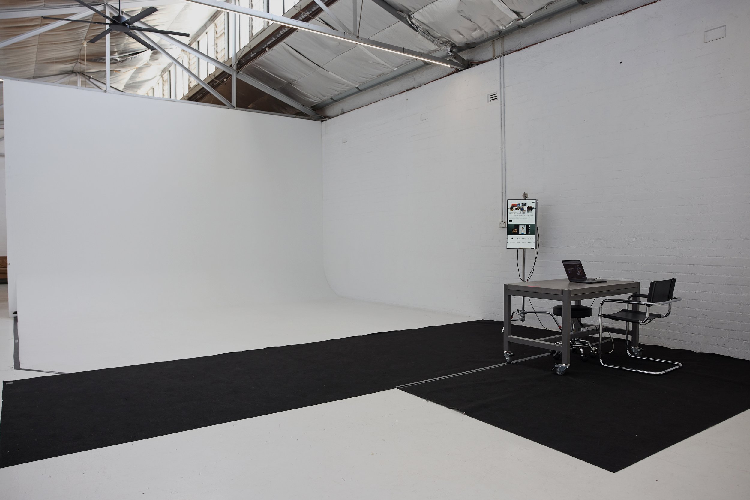 A large empty photography studio with a white wall backdrop, black carpeted floor, a table with a laptop, and a monitor mounted on the wall displaying a website.