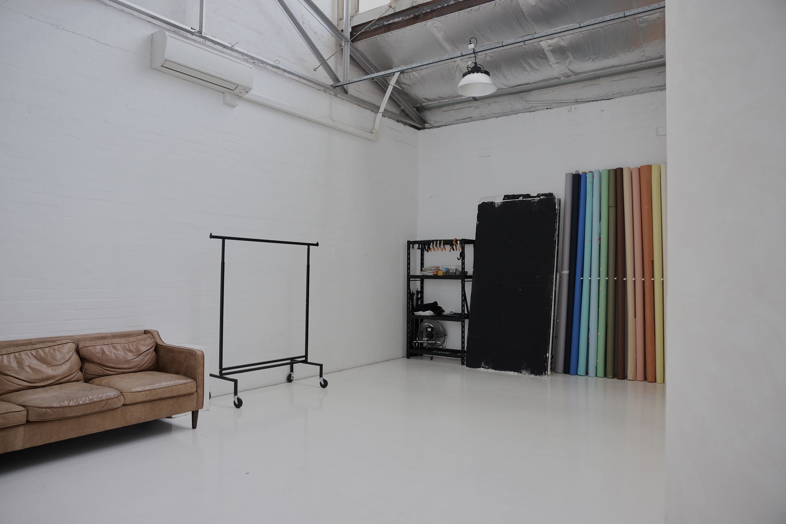 An art studio with a brown leather sofa on the left, a clothing rack on wheels, a black metal shelving unit, large blackboard, and colorful fabric or paper rolls against the white wall, with a high ceiling and bright lighting.