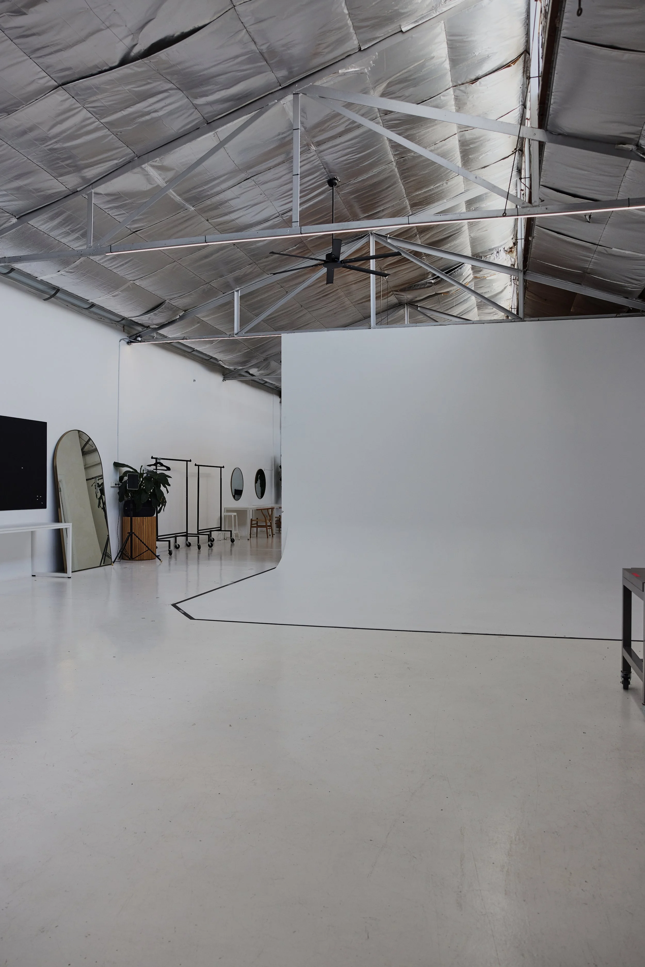 An empty photography studio with a white cyclorama wall, a black ceiling fan, a mirror, and various clothing racks and decor items against a white wall.