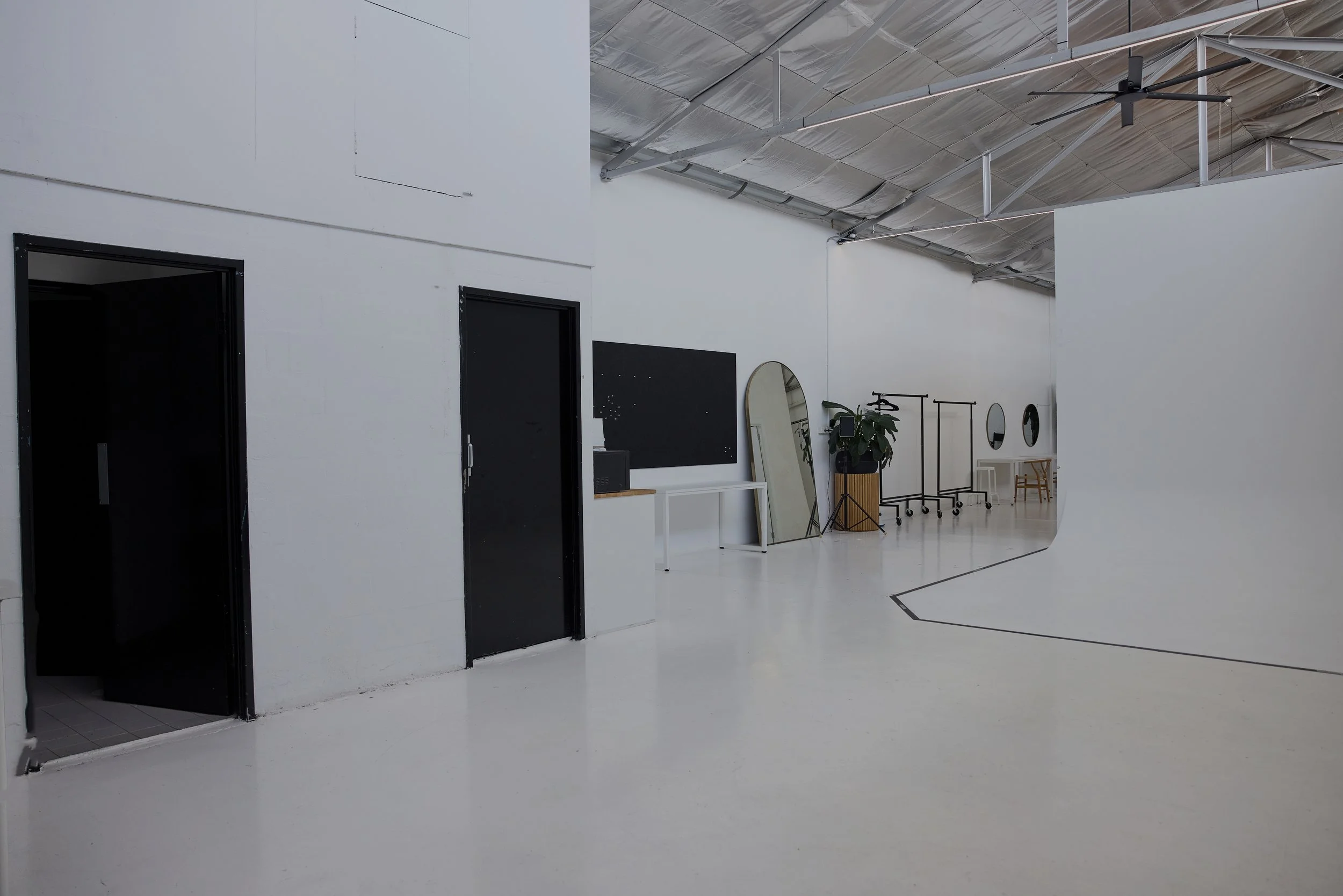 A minimalist photography studio with white walls, black doors, mirrors, and a high ceiling with visible metal beams and a ceiling fan.