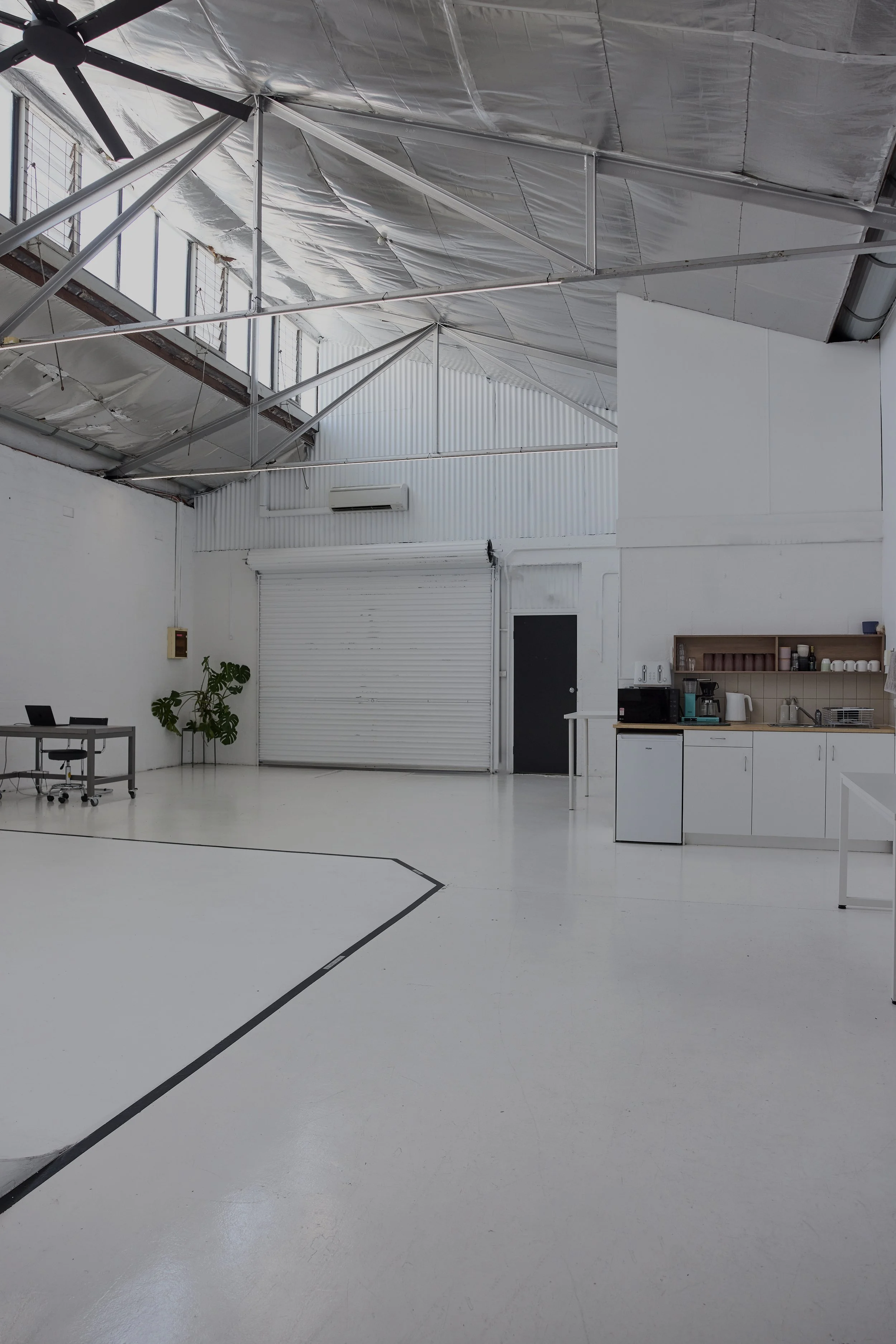 A spacious, industrial-style room with white walls, ceiling, and floor. The ceiling shows exposed metal beams and insulation. The right side of the room features a small kitchenette with a coffee machine, microwave, and open shelves. There is a black