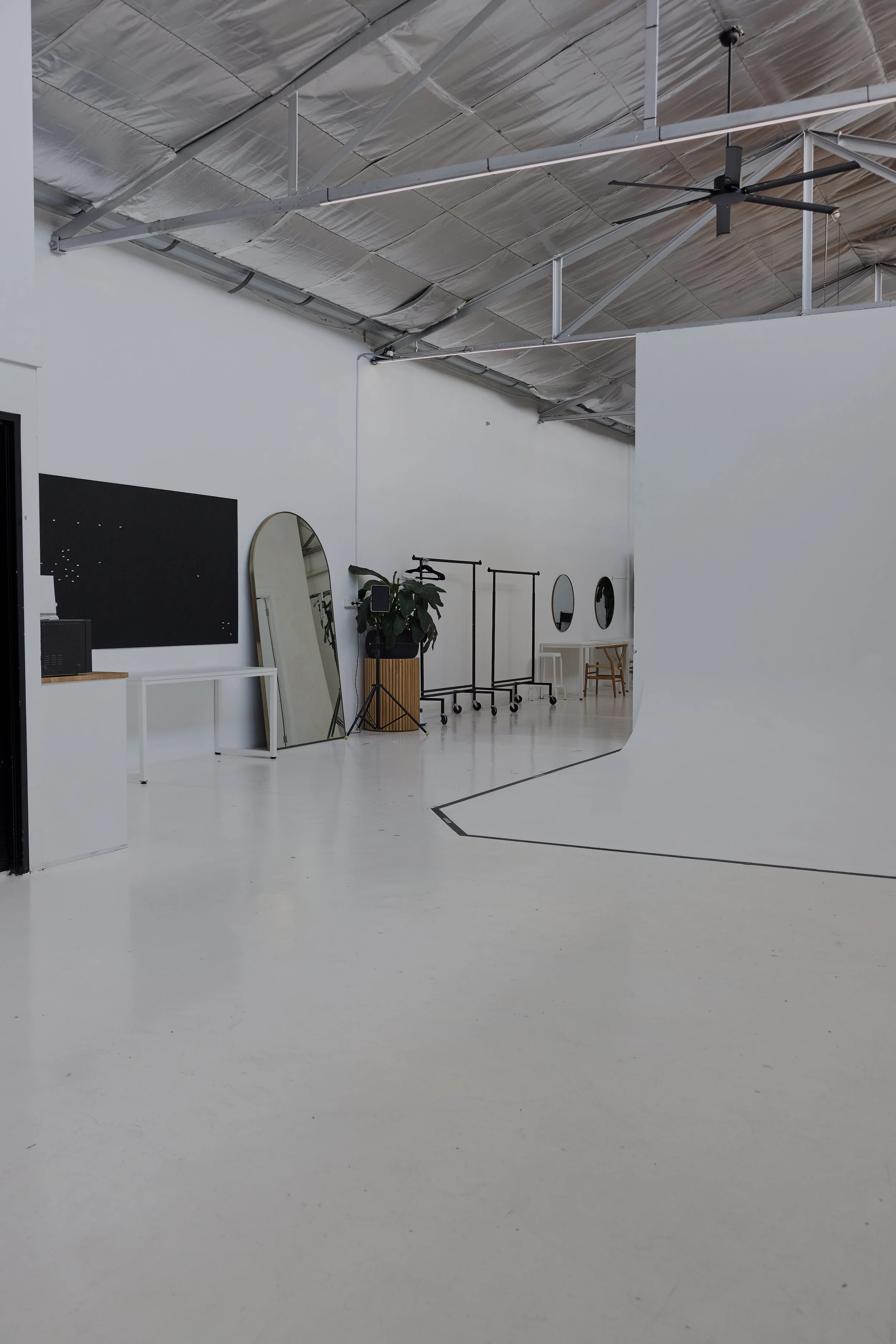 A modern, minimalist studio or workspace with white floors and walls, featuring large mirrors, a plant, clothing racks, and a white curved backdrop for photography or filming.
