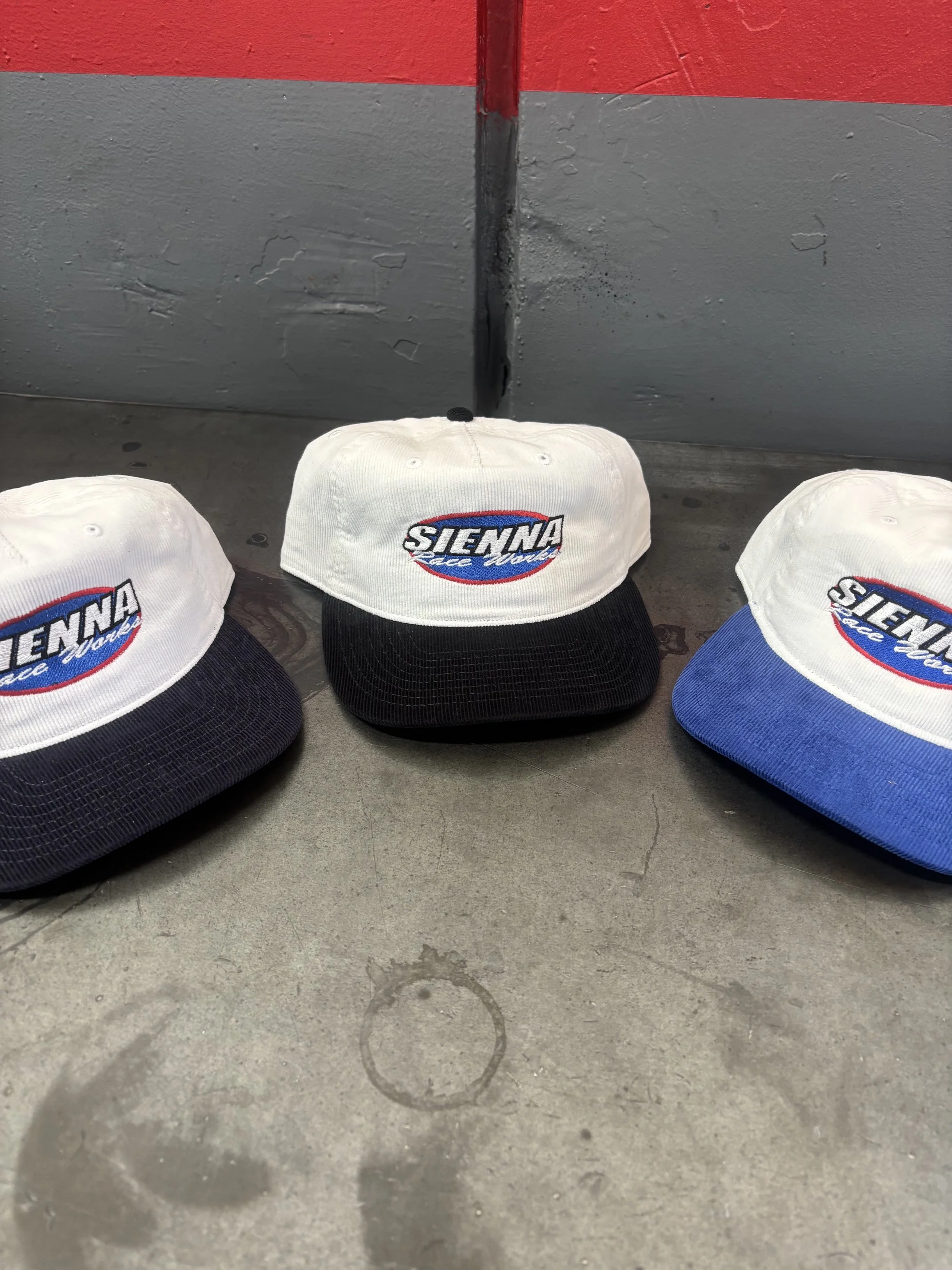 Three white Sienna Race Works branded hats on a gray concrete floor