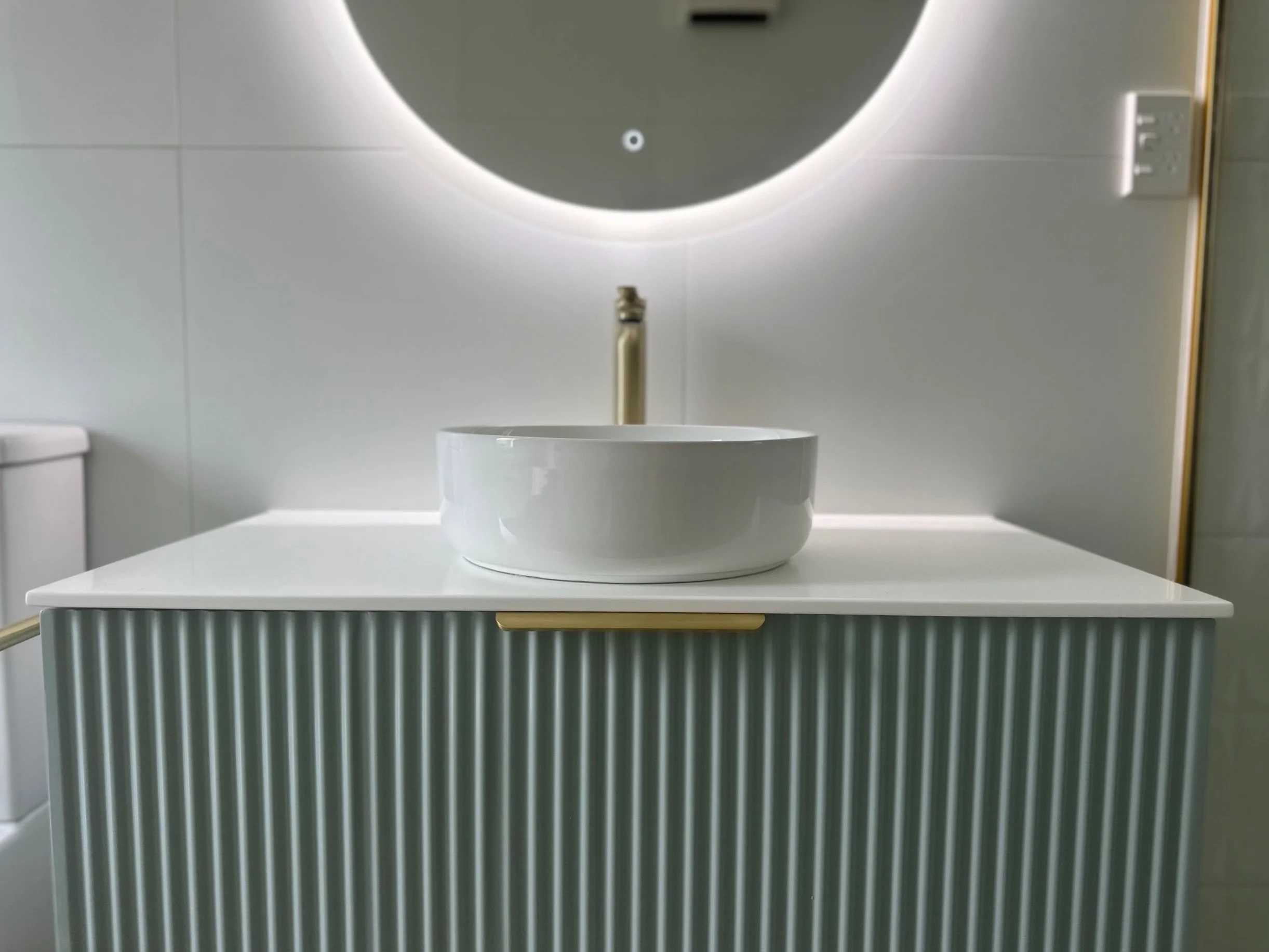 A modern bathroom sink with a white circular vessel basin on a white countertop, positioned on a turquoise ribbed vanity, with a large backlit mirror and a gold towel bar.