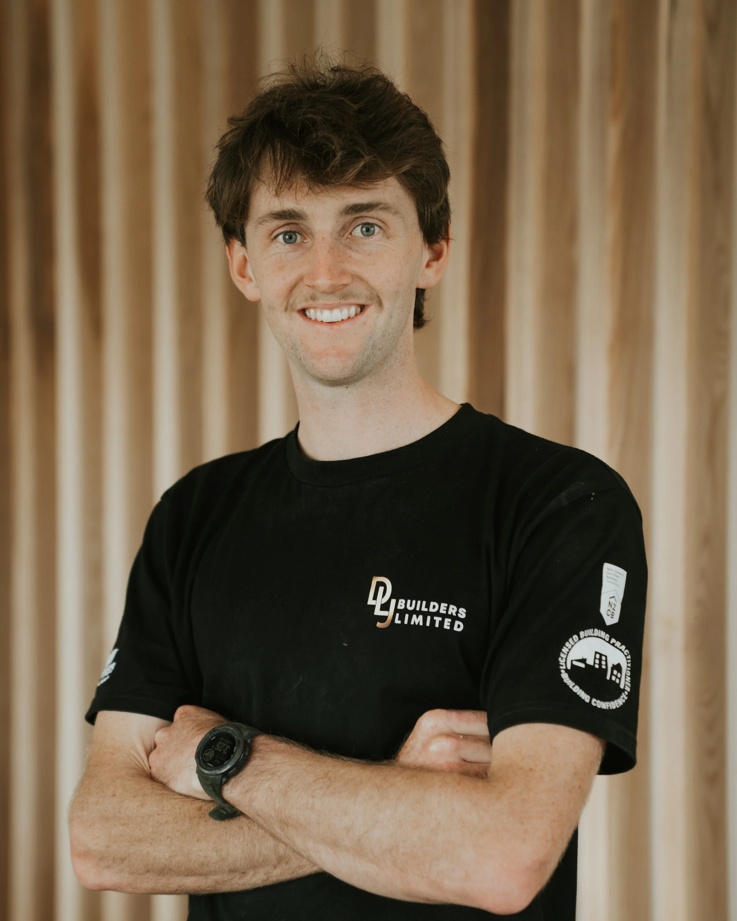Young man with short brown hair smiling, standing with arms crossed, wearing a black hoodie with a company logo, in front of a wooden construction framework.