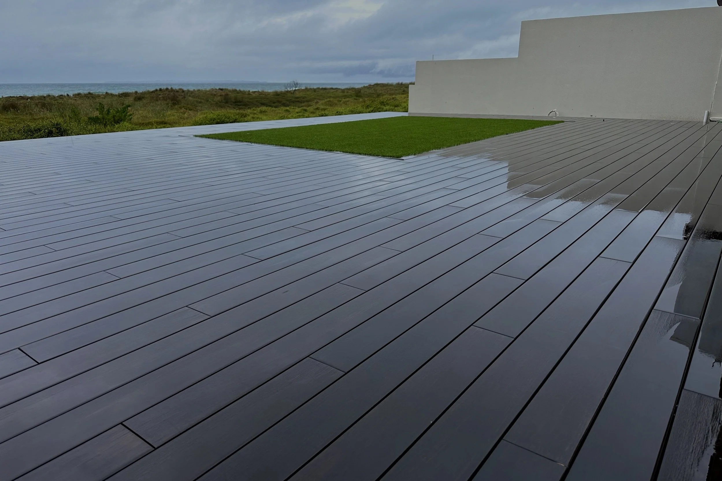A wet, dark wooden deck with a small patch of green grass, overlooking a landscape with bushes and an ocean in the distance under an overcast sky.