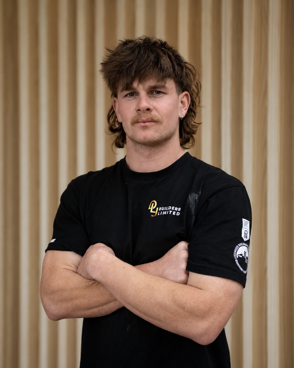 A young man with brown, tousled hair and a mustache, wearing a black T-shirt with a logo and text, standing indoors in front of a wooden wall with windows, crossing his arms and looking at the camera.