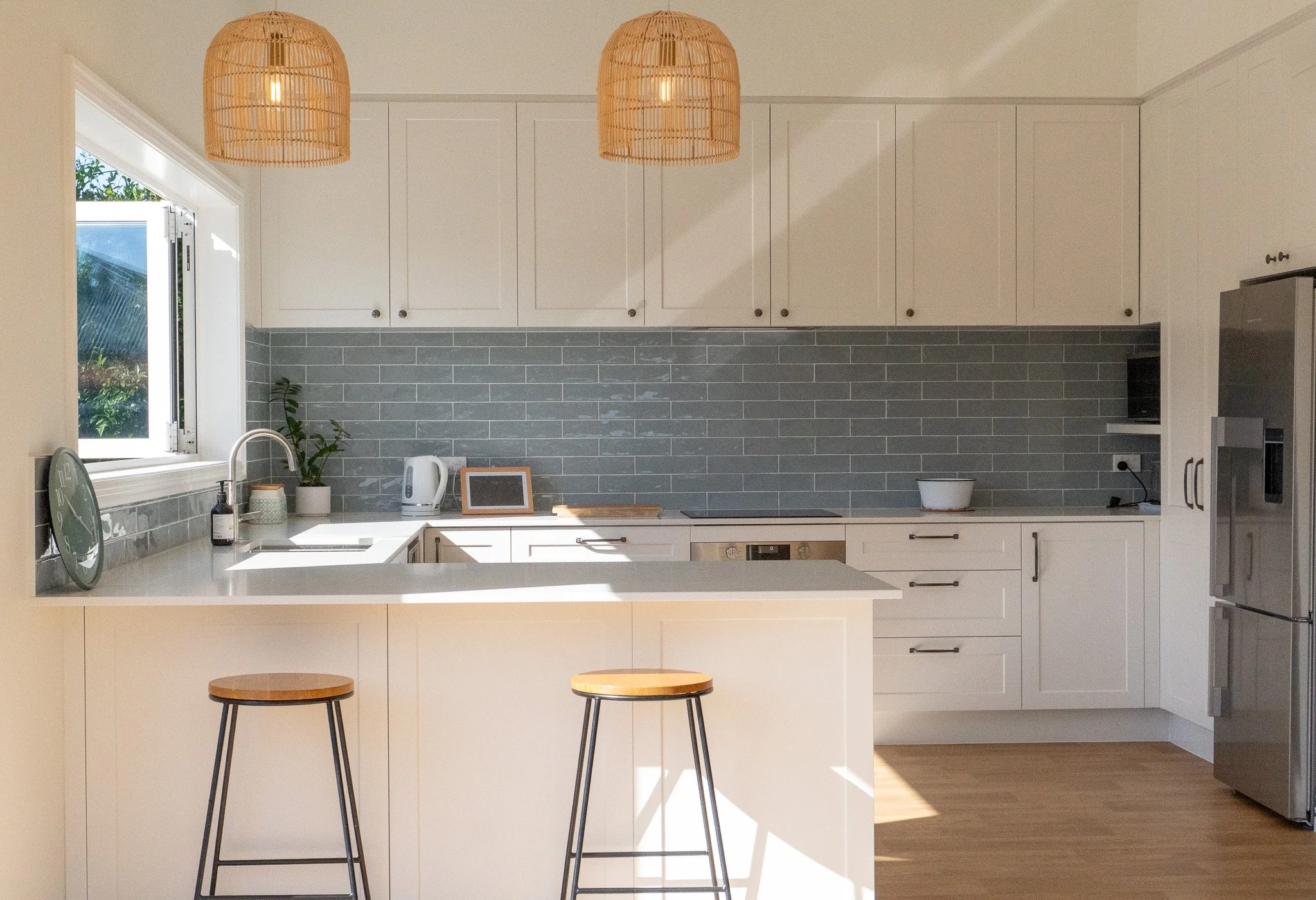 Modern kitchen with white cabinets, gray backsplash tiles, a window with sunlight, a white kitchen island with two wooden stools, and stainless steel appliances.