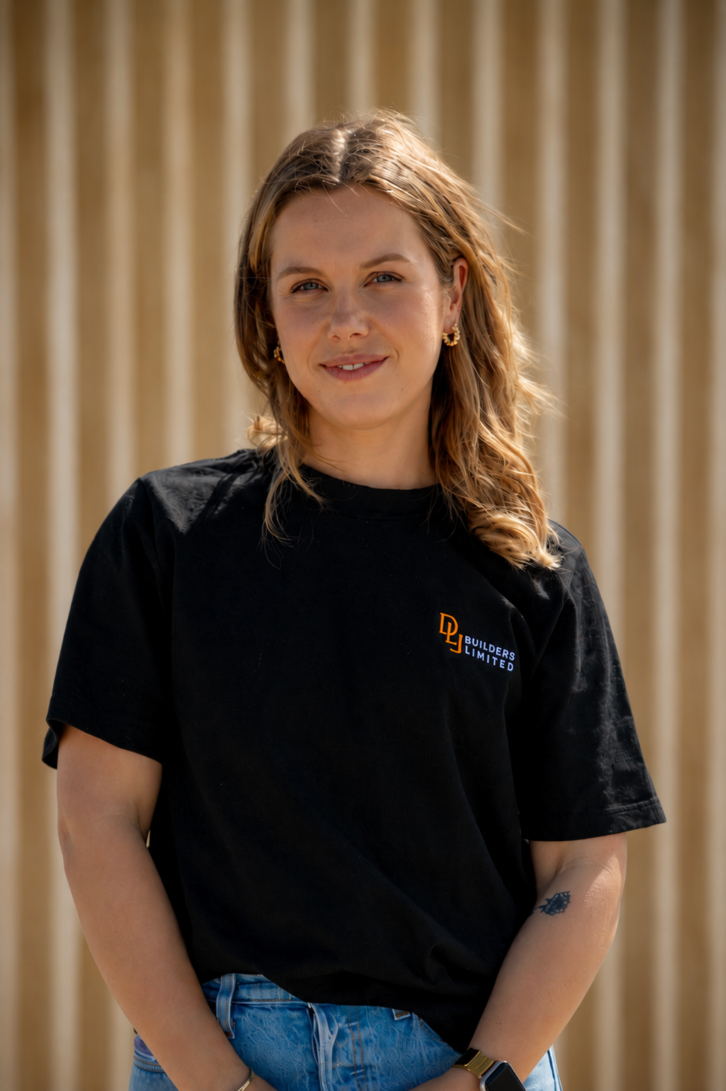 A woman standing on a sandy beach with a mountain in the background. She has light brown hair, is wearing a black T-shirt with a logo on it, blue jeans, and a smartwatch. The sky is partly cloudy.