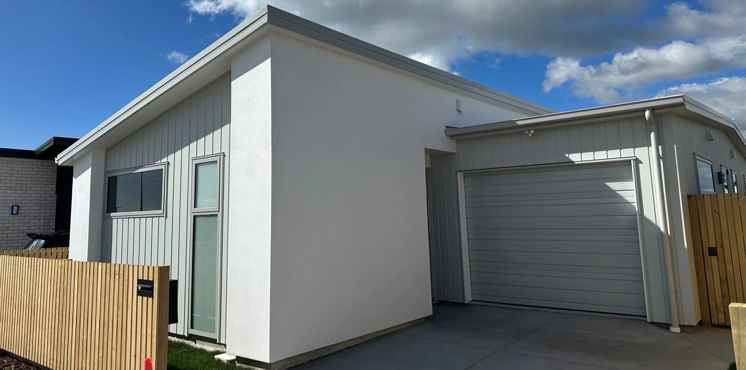 Modern house with white exterior walls, grey metal siding, a garage door, a wooden fence, and a concrete driveway under a partly cloudy sky.