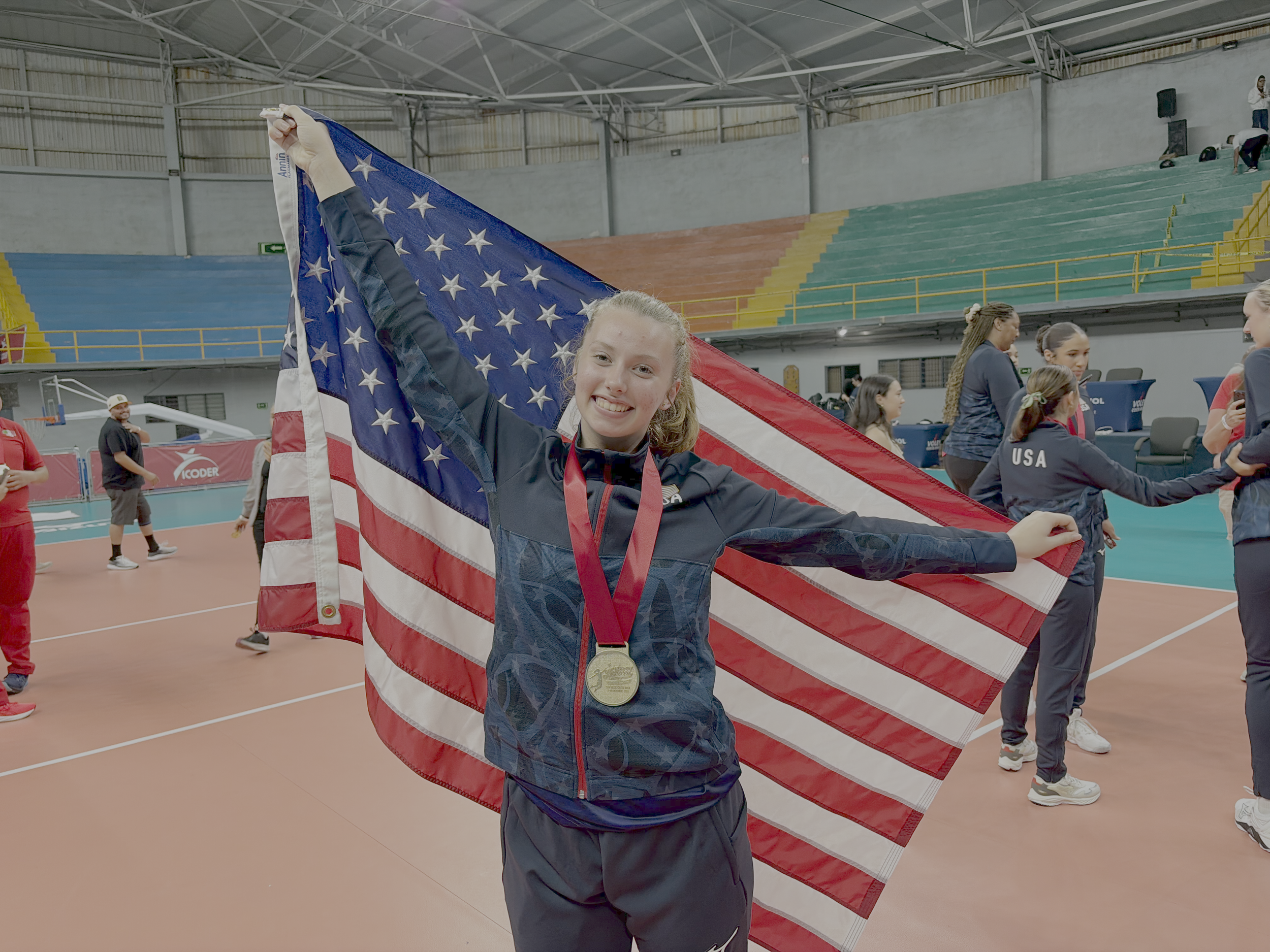 A young female athlete smiling, holding an American flag and wearing a medal around her neck in an indoor sports arena, with other athletes in the background.