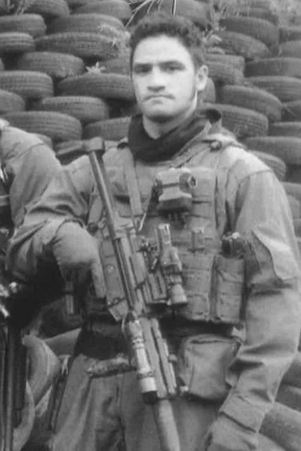 A man in military gear holding a rifle, standing in front of stacked tires.