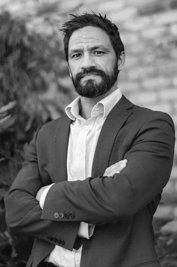 A man with a beard and mustache, wearing a suit jacket and white dress shirt, standing outdoors with arms crossed and looking at the camera.