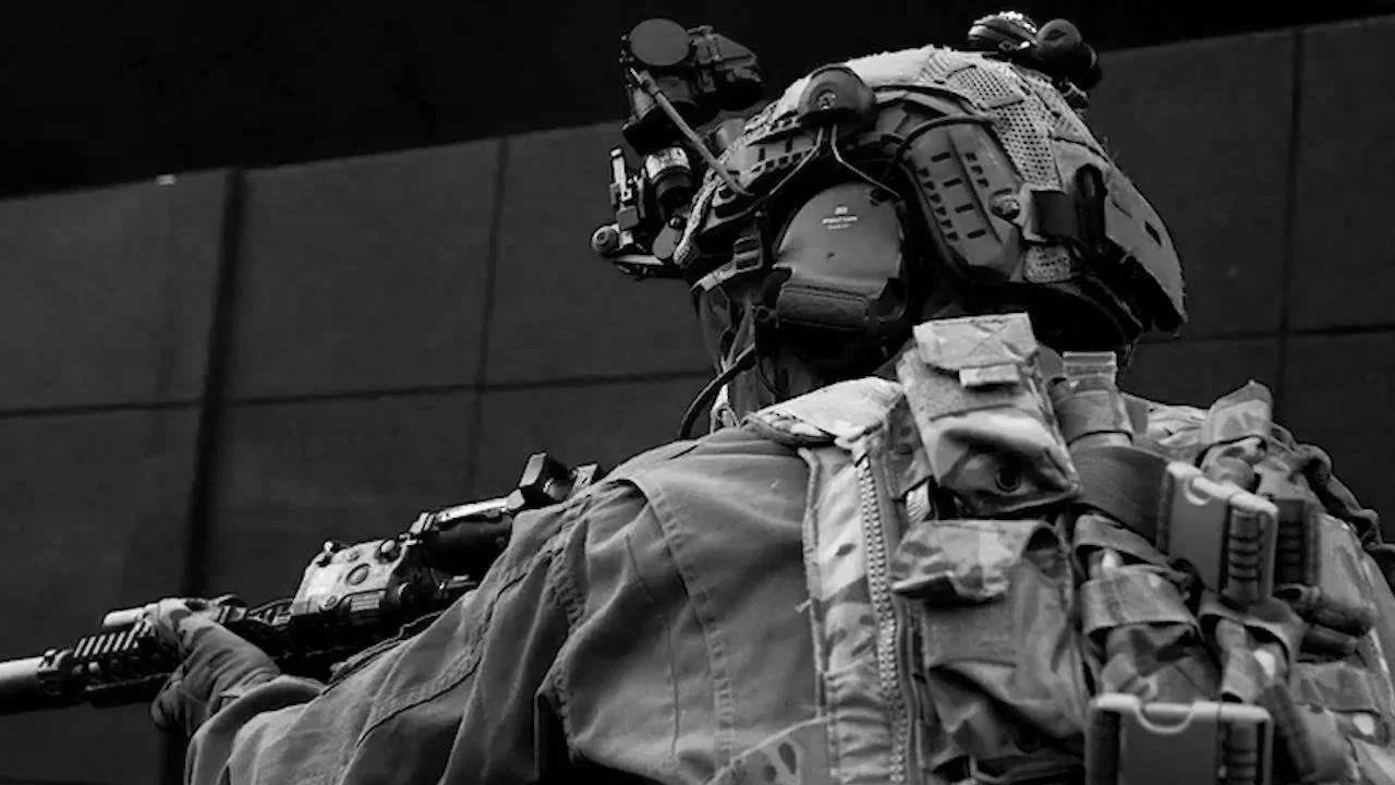 Close-up of a soldier in tactical gear carrying a rifle, wearing a helmet with attachments, and standing against a dark, textured background.