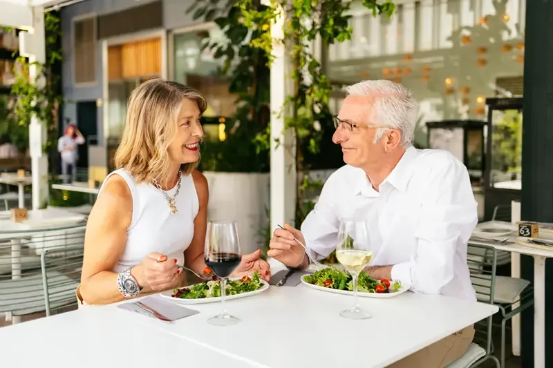 a man and a woman sitting at an outdoor restaurant, each with a salad and a glass of wine. They are looking towards each other, smiling.