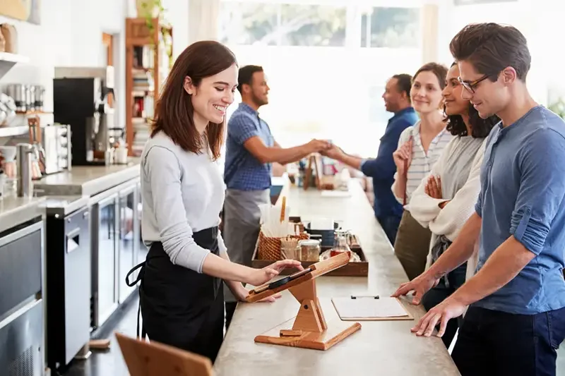 A lady serving a man in at a cafe counter. She is taking down his order on a tablet housed in a wooden stand. There are people smiling in the background, waiting to order.