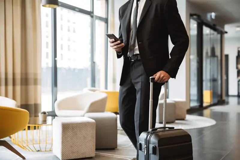 man in a suit wheeling a suitcase through a hotel lobby. He is holding a phone in the other hand