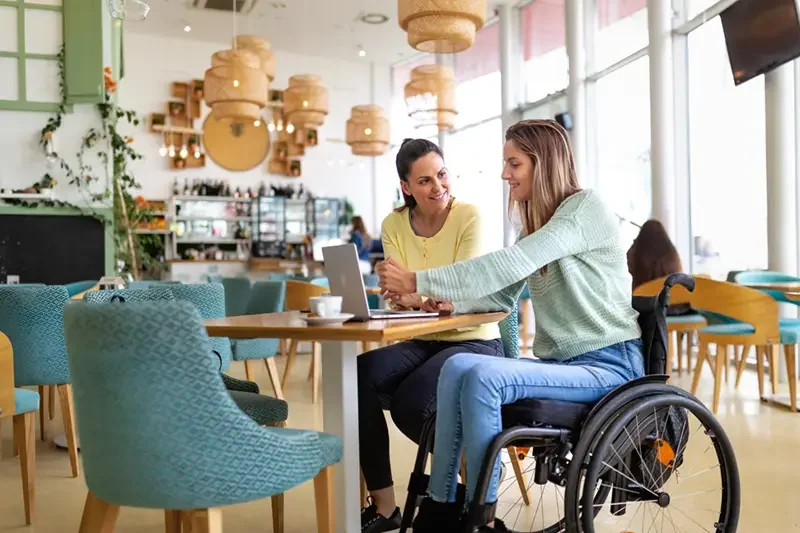 two ladies sitting at a table in a cafe. one is wearing a light green jumpers with blue jeans and is sitting in a wheelchair. The lady next to her is in a bright yellow long-sleeve shirt and dark pants, looking towards the first lady, smiling.