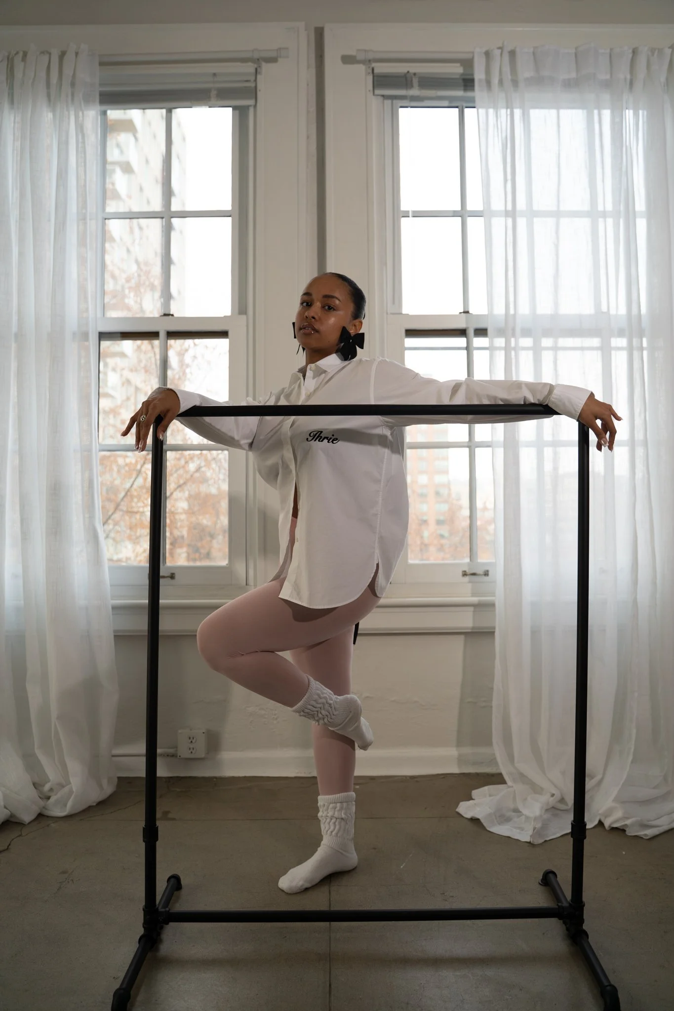A woman in a white shirt and pale pink leggings practicing ballet en pointe inside a room with large windows and sheer white curtains. She is holding a ballet barre and standing on one foot with her other foot raised behind her.