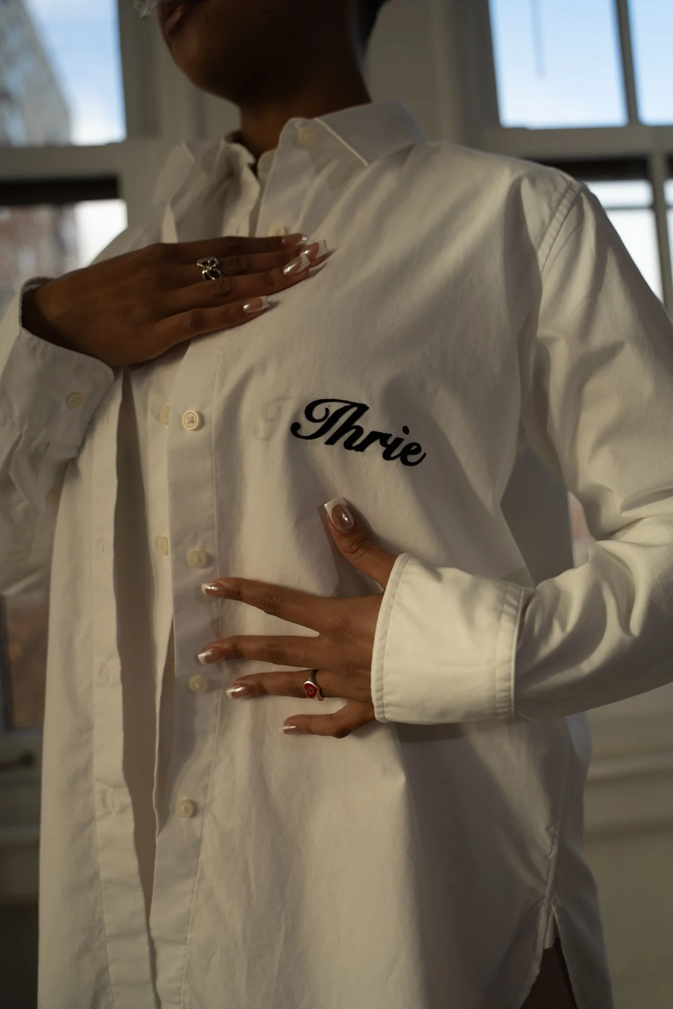 Person wearing a white shirt with the name "Thrie" embroidered on it, standing indoors near windows, with one hand on their chest and the other on their stomach.