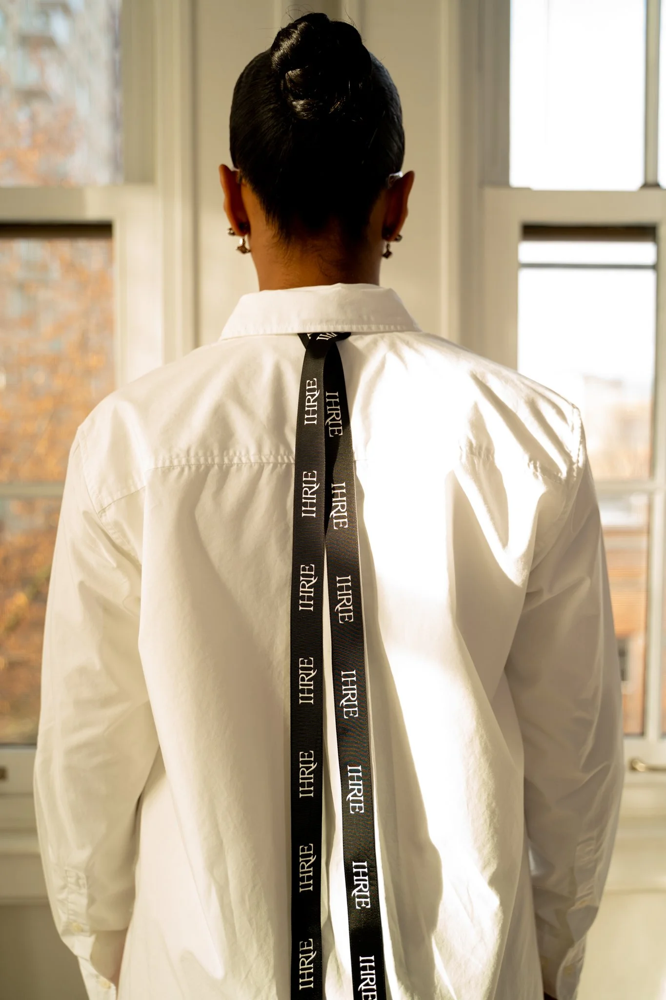 Back of a woman with dark hair in a bun, wearing a white shirt, standing in front of windows with sunlight shining through, with black ribbons adorned with the word 'THREE' hanging from her neck.